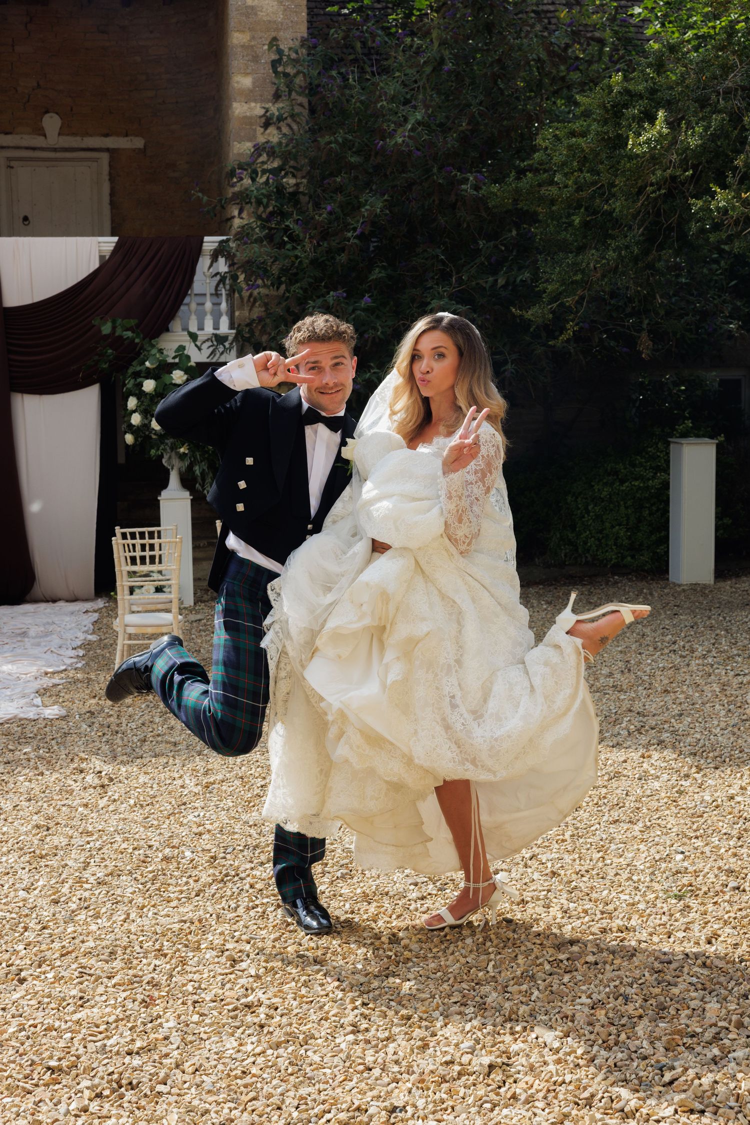 Playful groom in tartan kilt carries laughing bride in voluminous white dress at outdoor wedding.Greater Manchester