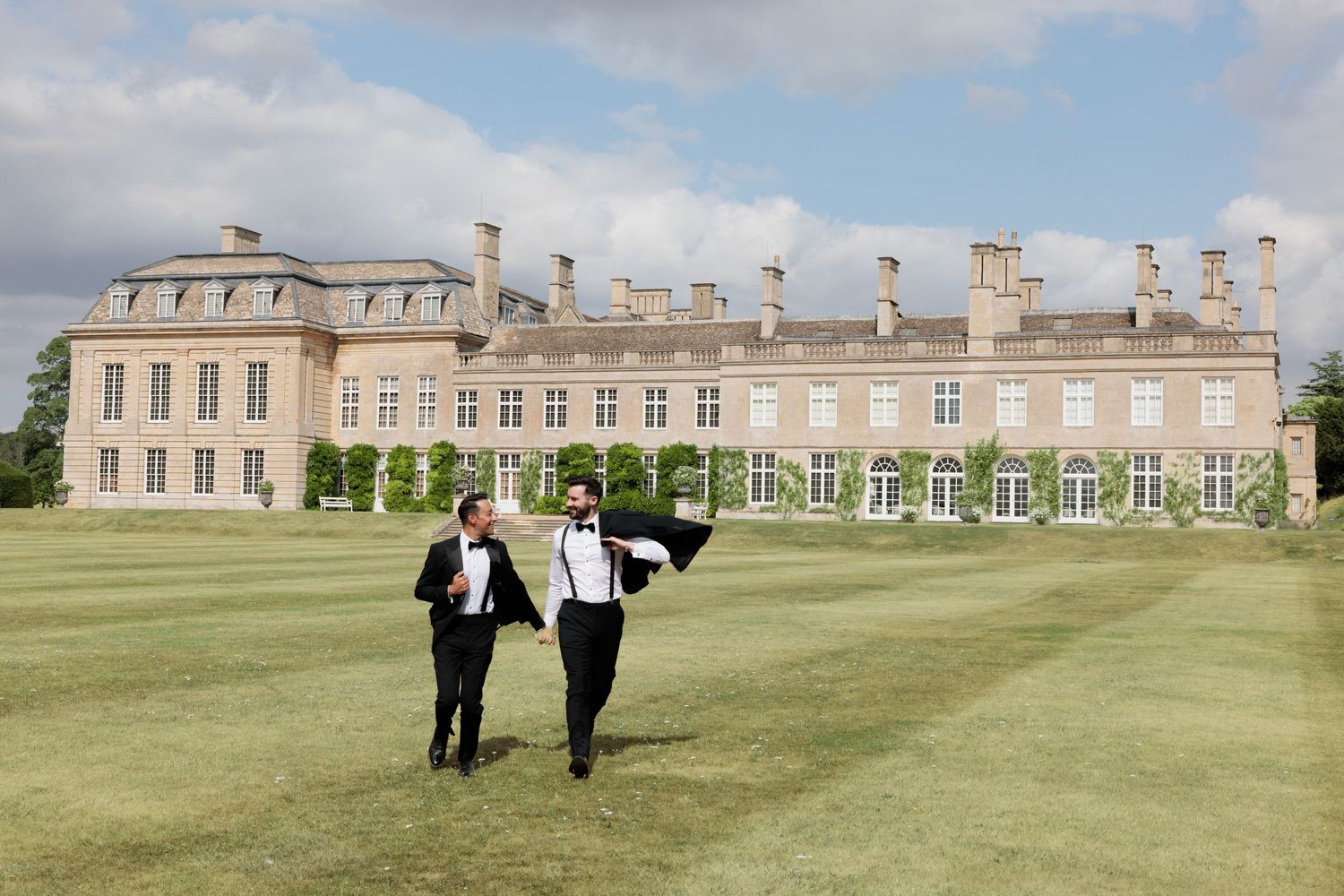 Two grooms in black tuxedos walking across a vast green lawn in front of a grand English manor house.