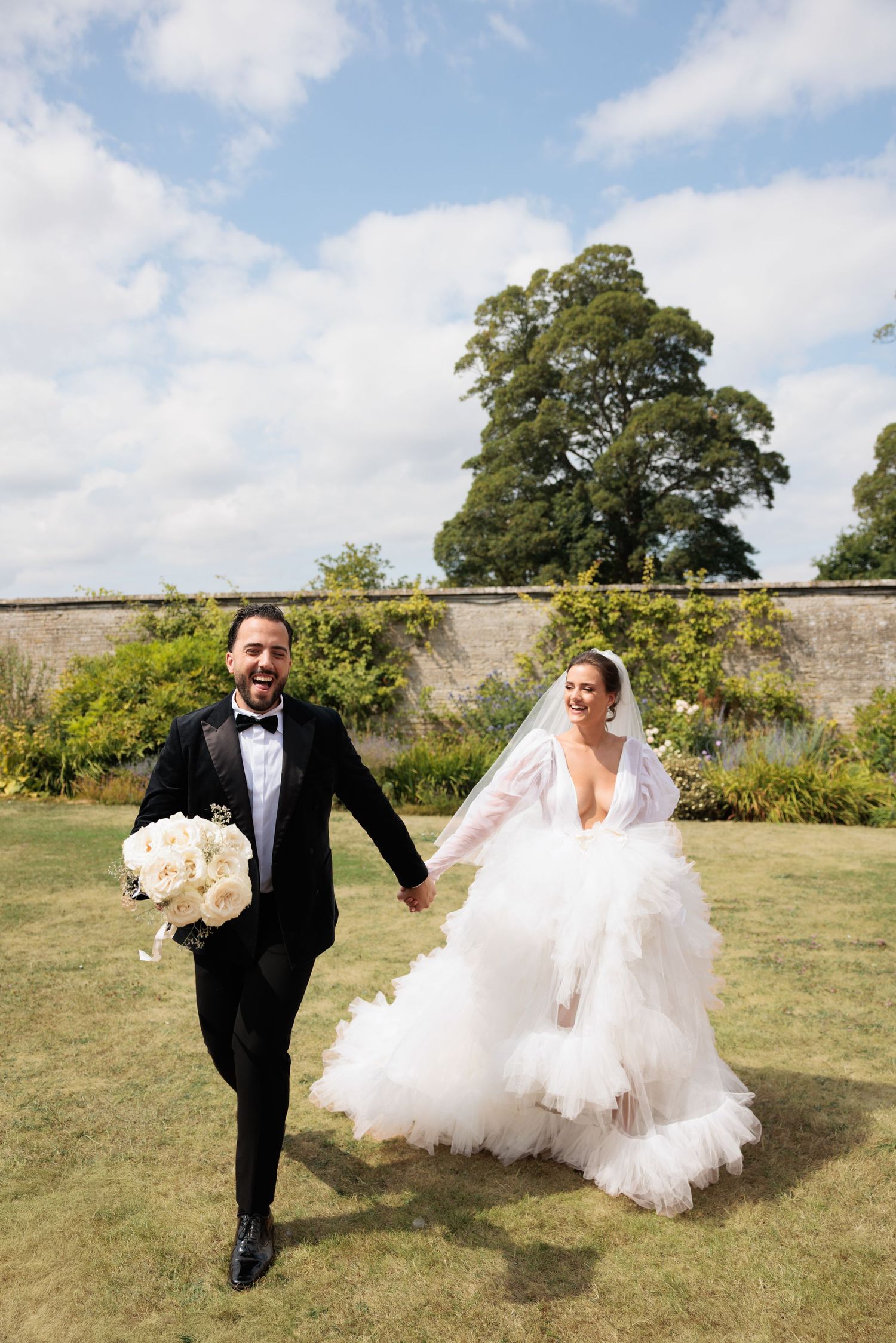 Laughing groom in black tuxedo holds white bouquet while running hand-in-hand with bride in ruffled gown outdoors.