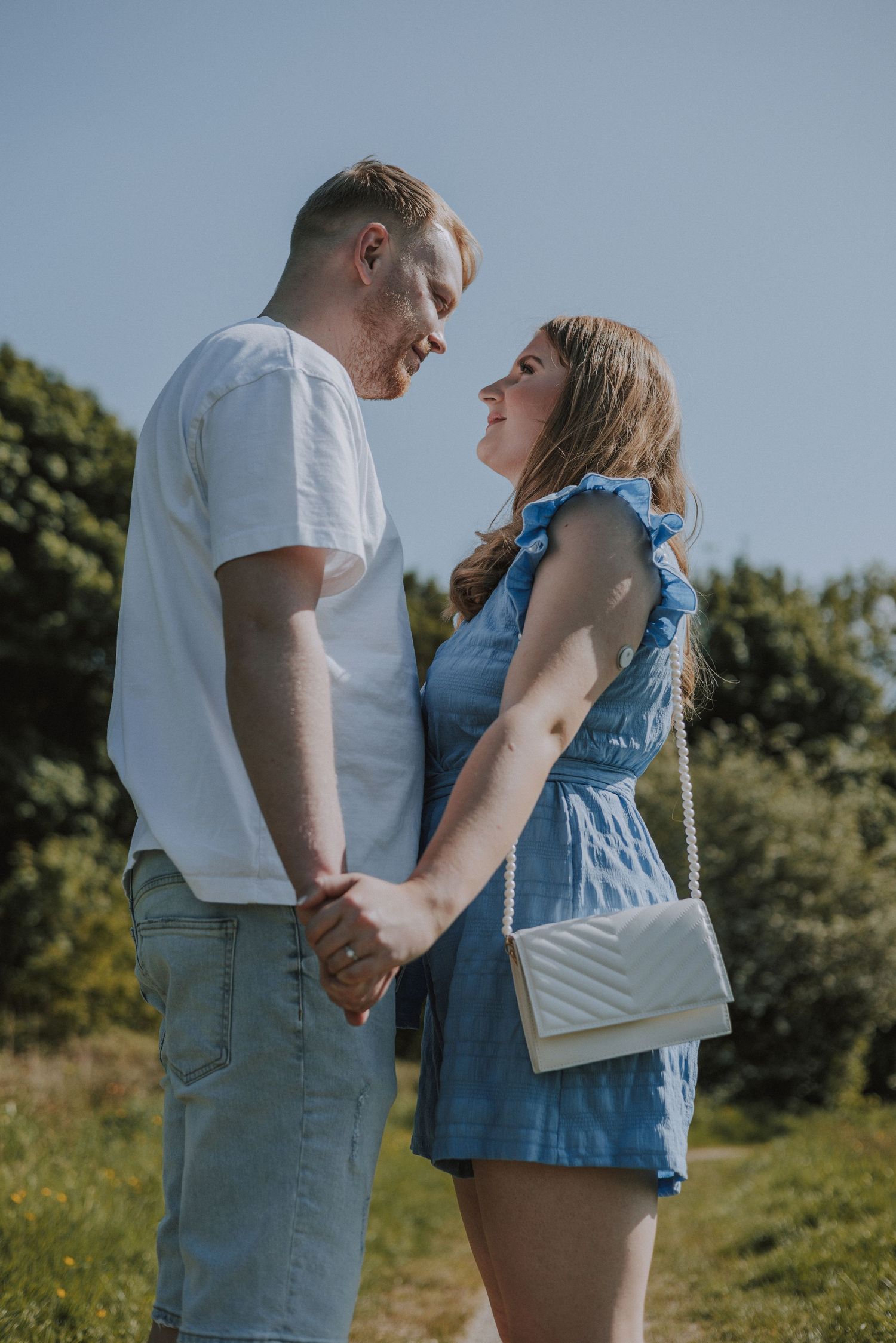 Young couple holding hands and smiling at each other on a sunny outdoor path, woman wearing blue dress with white purse. Haigh Hall Wigan UK