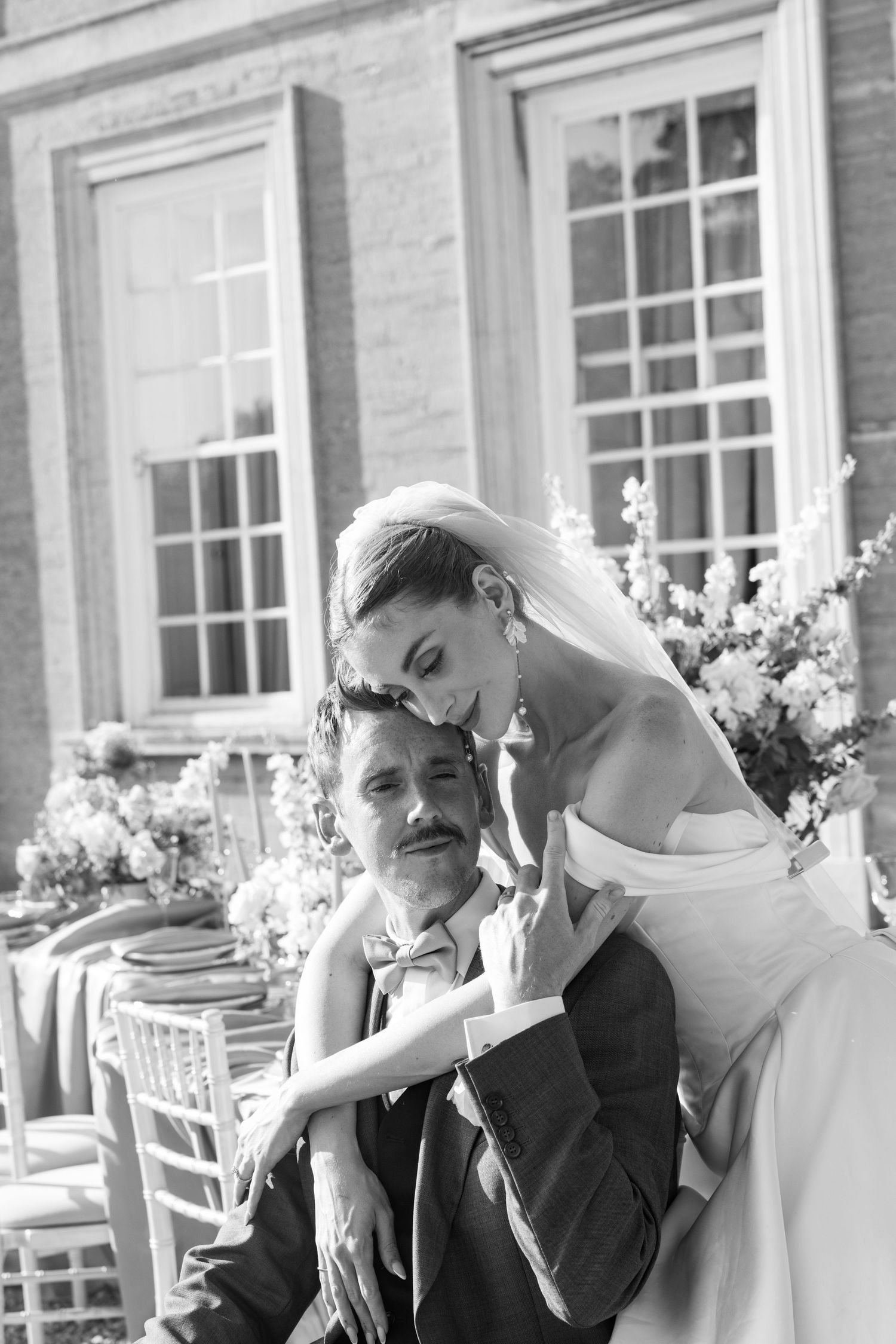 Black and white portrait of bride embracing seated groom at elegant outdoor wedding reception.
