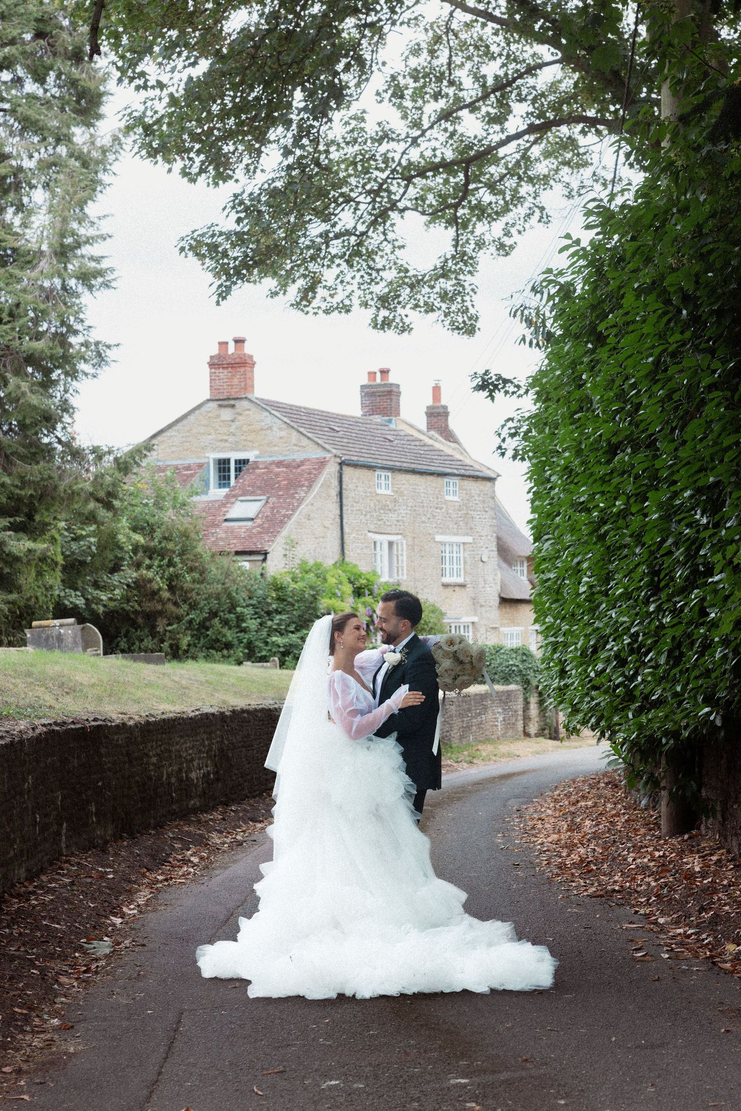 Bride and groom embrace on winding path surrounded by lush trees near charming countryside home.