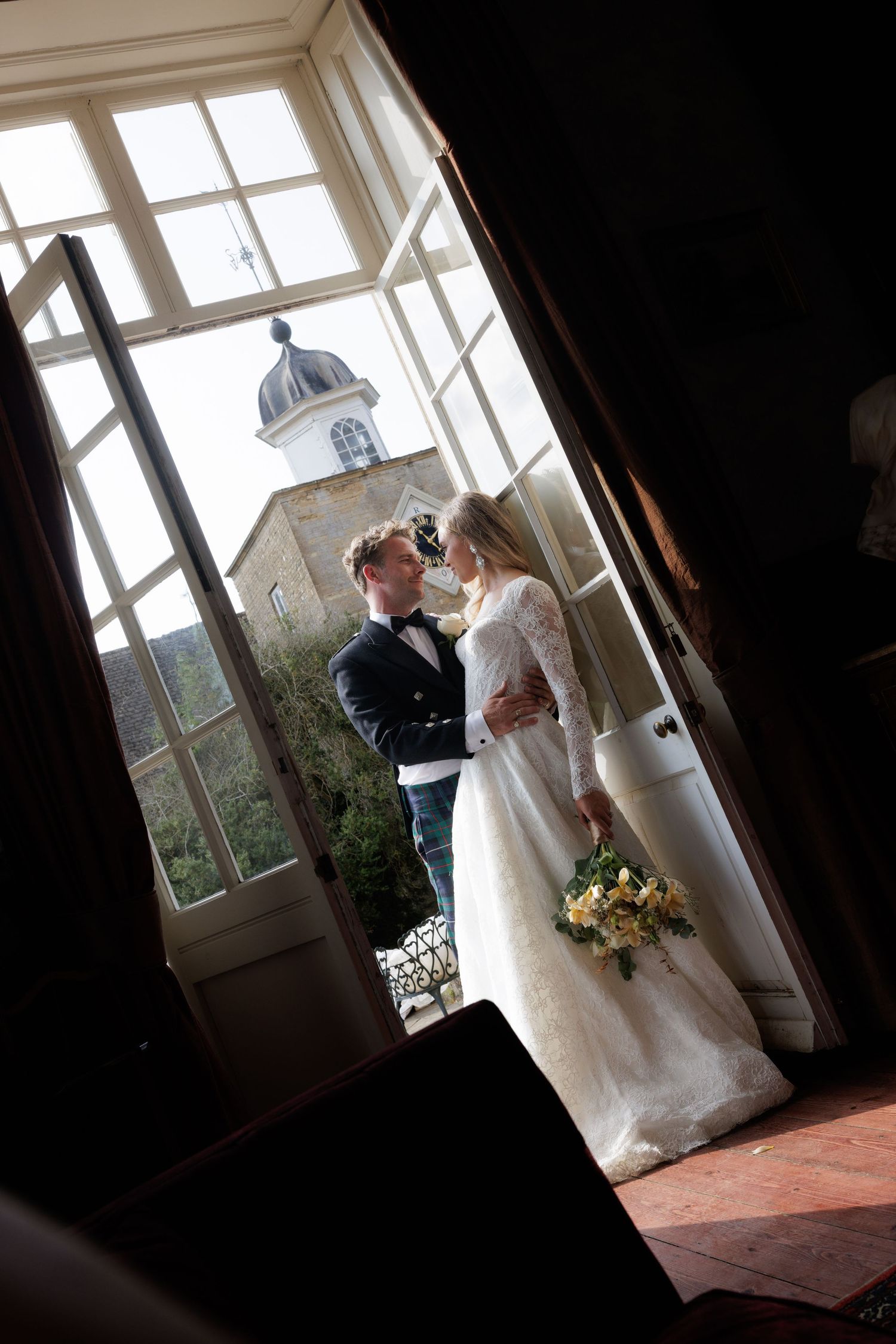 Bride and groom sharing an intimate moment on a balcony framed by elegant French doors with a church tower behind.