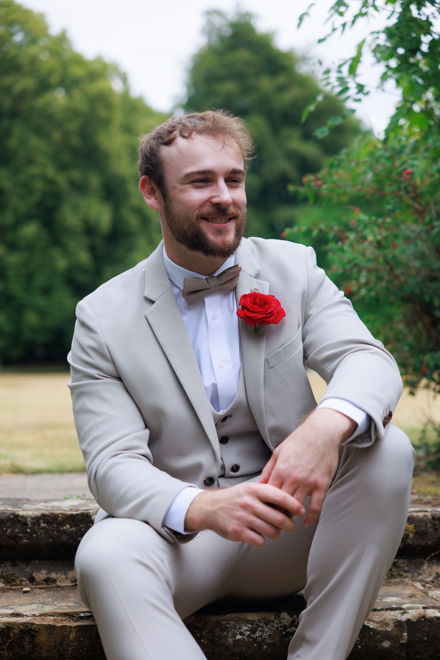 Smiling groom in a beige suit with red rose boutonniere and bow tie sitting relaxed on stone steps outdoors.