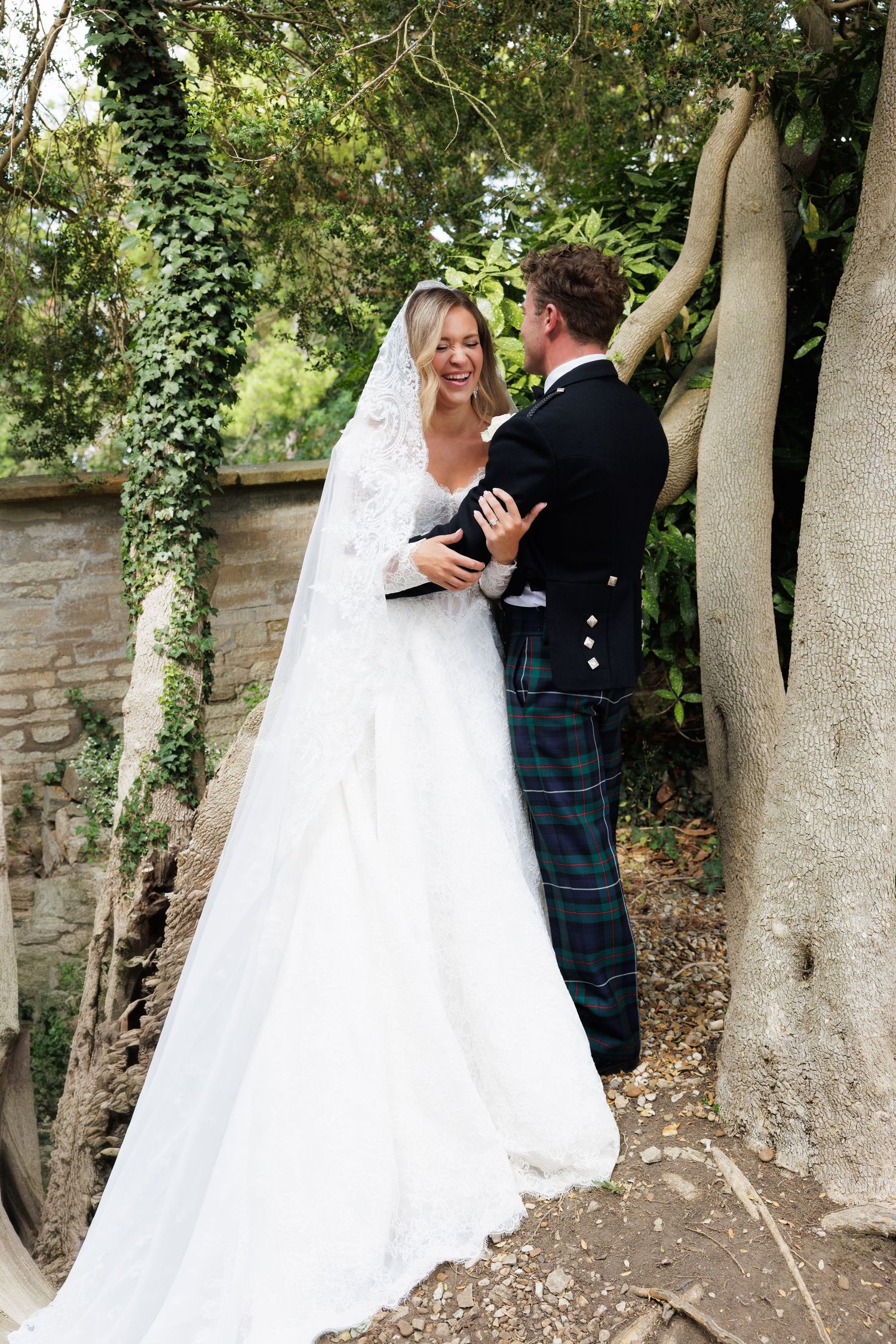 Bride in lace veil and groom in tartan kilt share romantic moment among ivy-covered trees. Wigan photographer