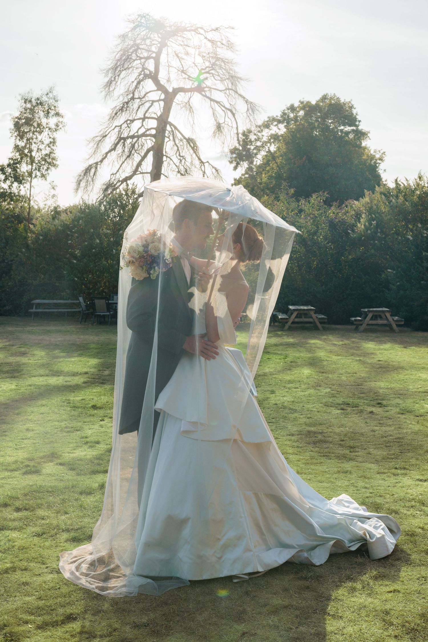 Bride and groom share intimate kiss beneath flowing veil in sunlit garden at golden hour.