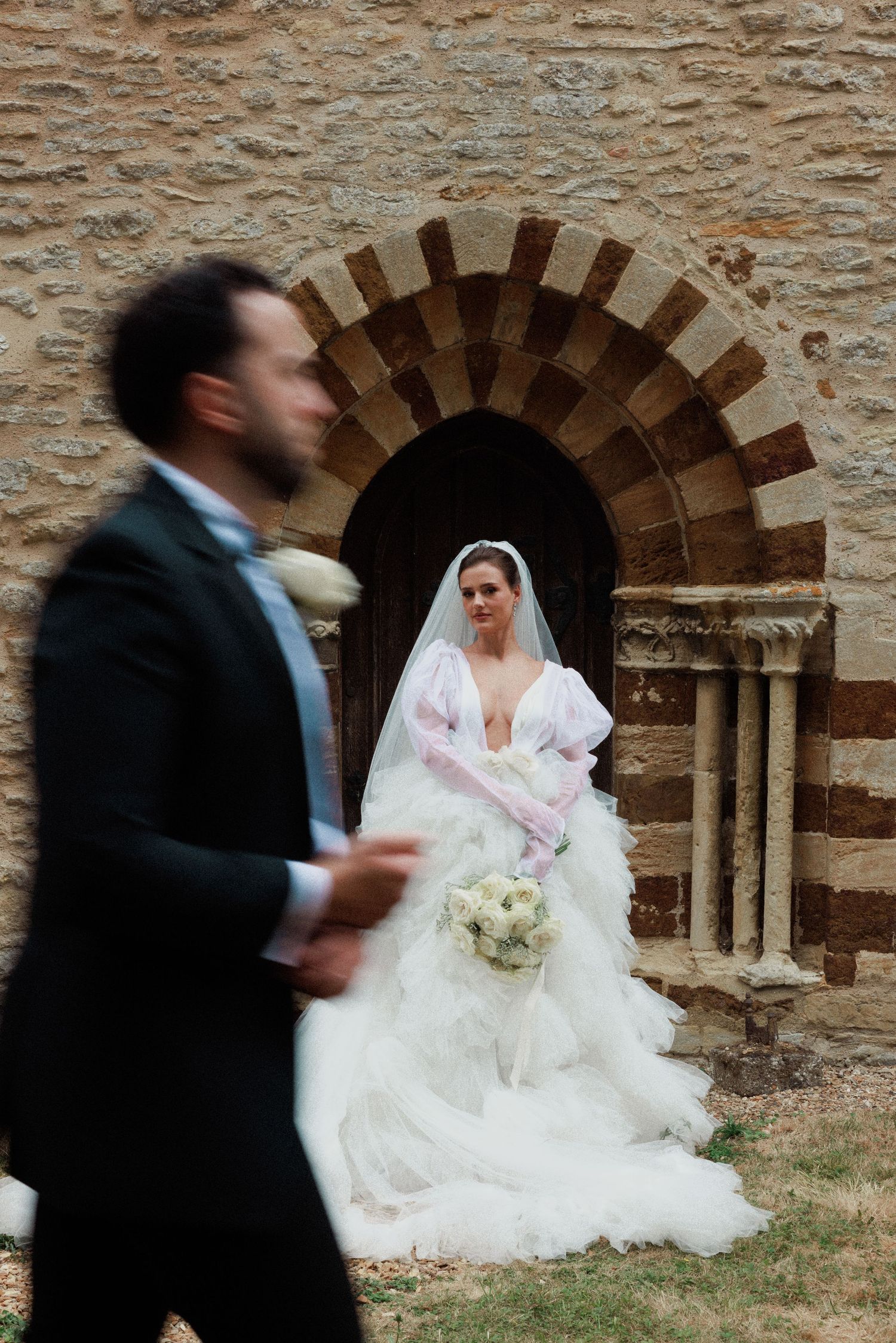 Bride in white gown stands in stone archway as groom walks past in blur at outdoor wedding.