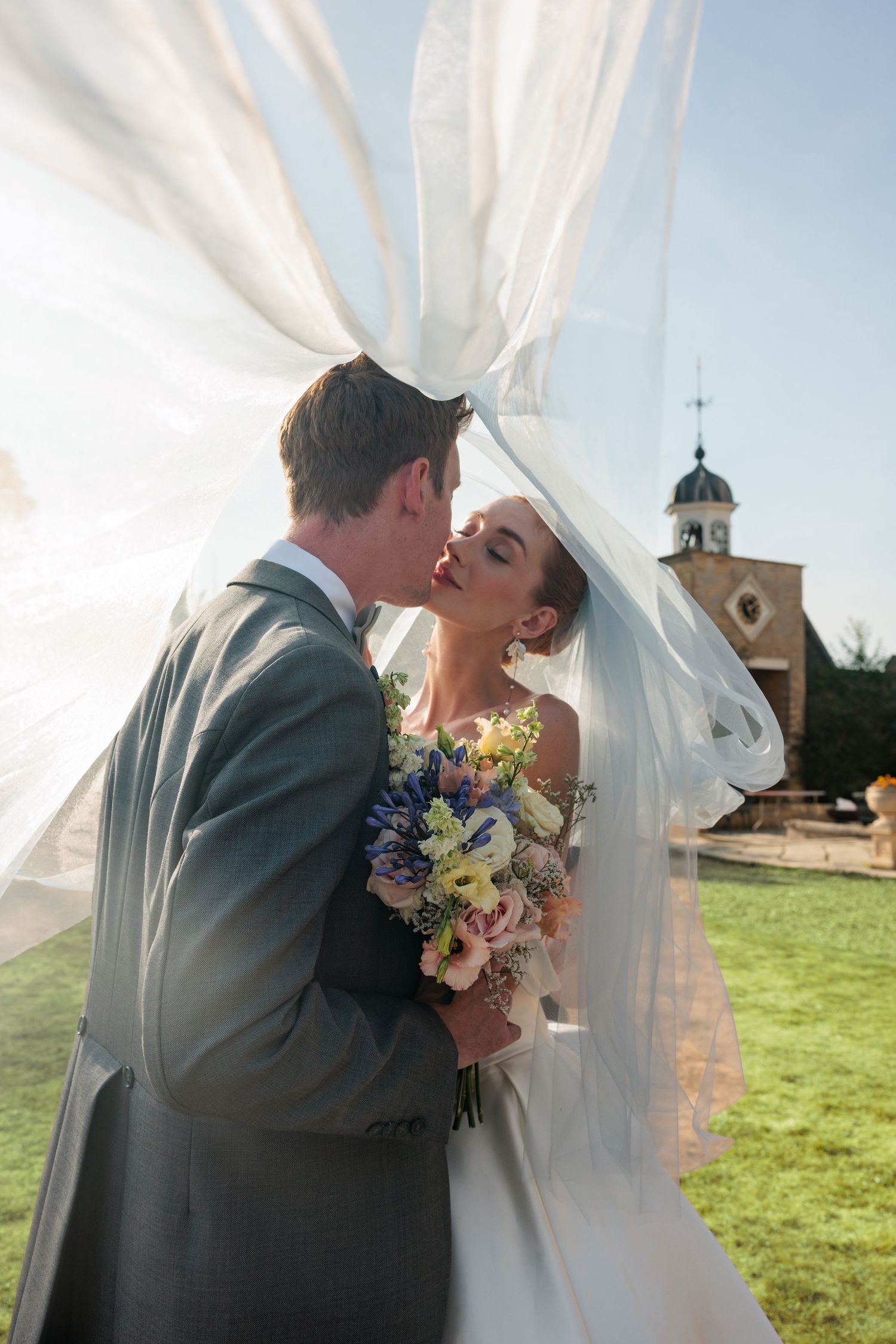 Bride and groom sharing a romantic kiss under flowing white fabric drapes outdoors on their wedding day.
