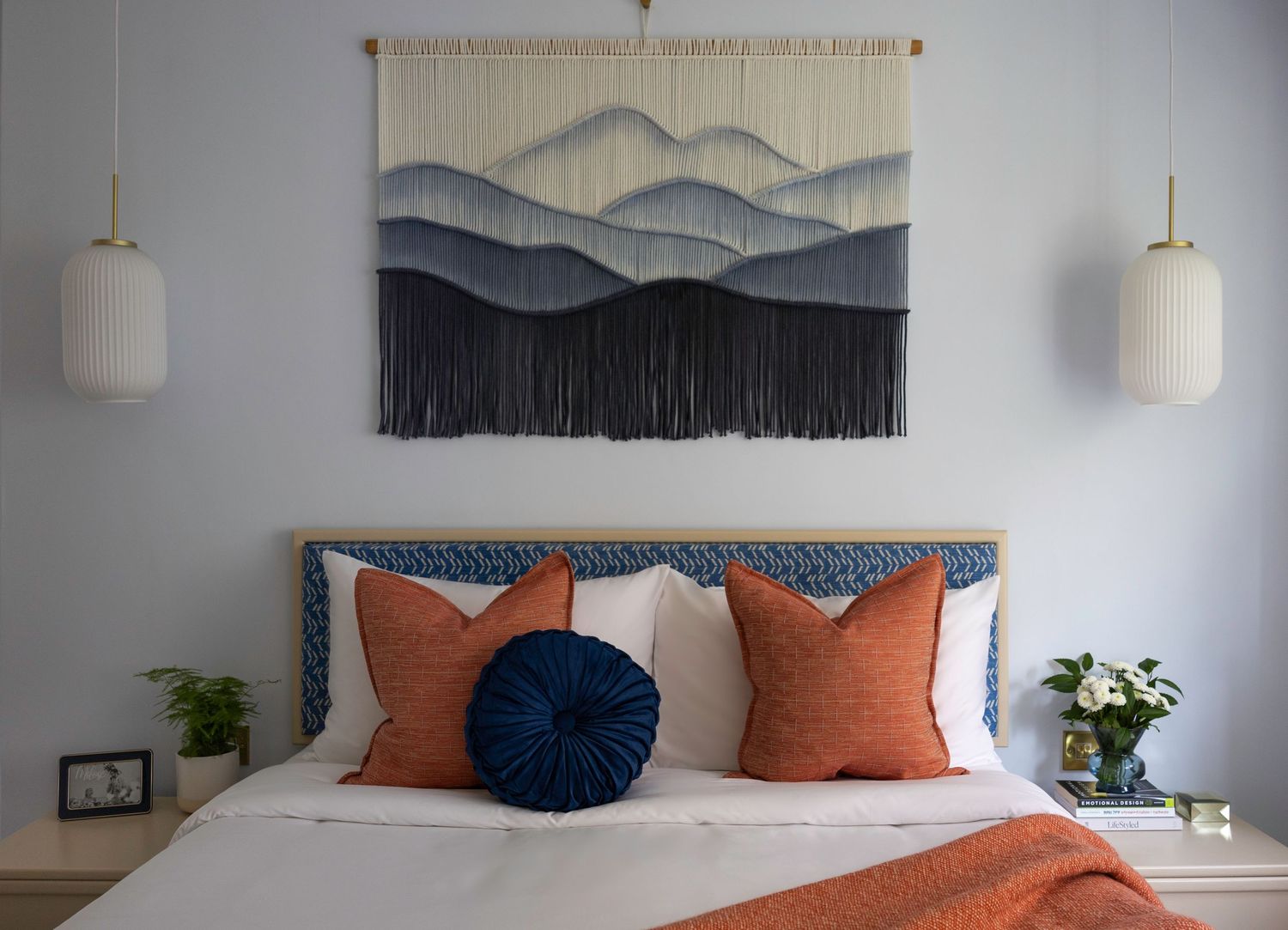 Bedroom with blue ombre macrame wall hanging above blue upholstered bed with orange pillows.
