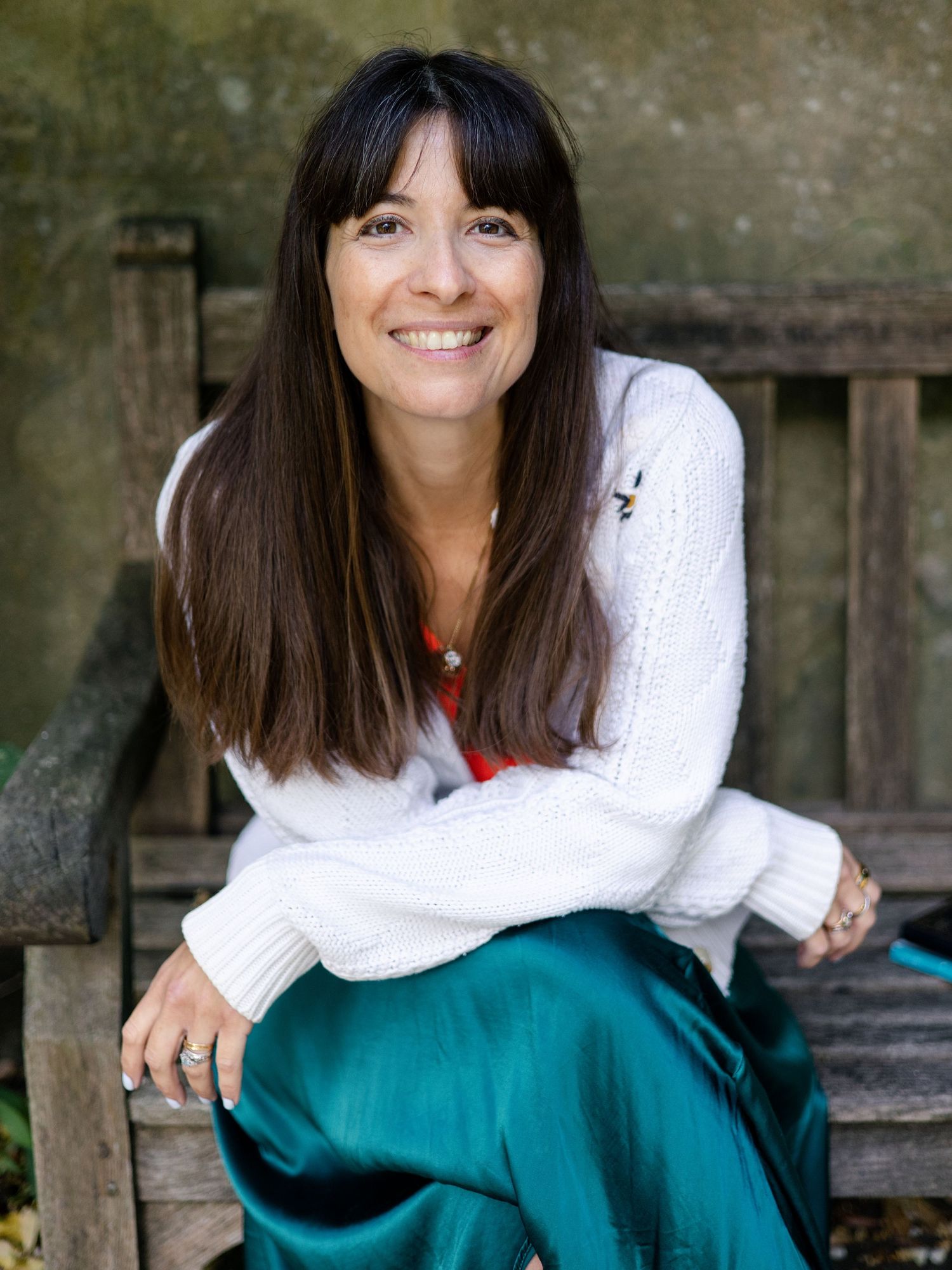 Smiling woman with dark hair in white sweater and teal satin skirt sitting on wooden bench outdoors.