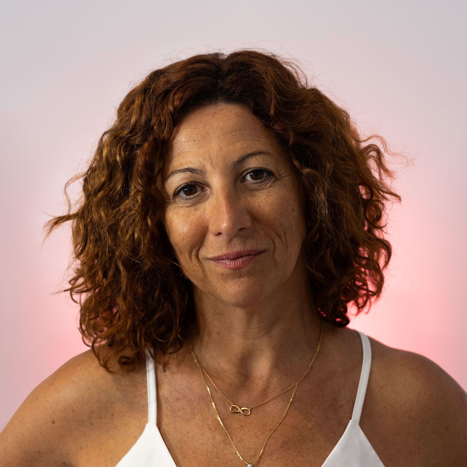 Woman with curly auburn hair wearing a white top and gold necklaces against a pink background. Lloret de Mar, Spain