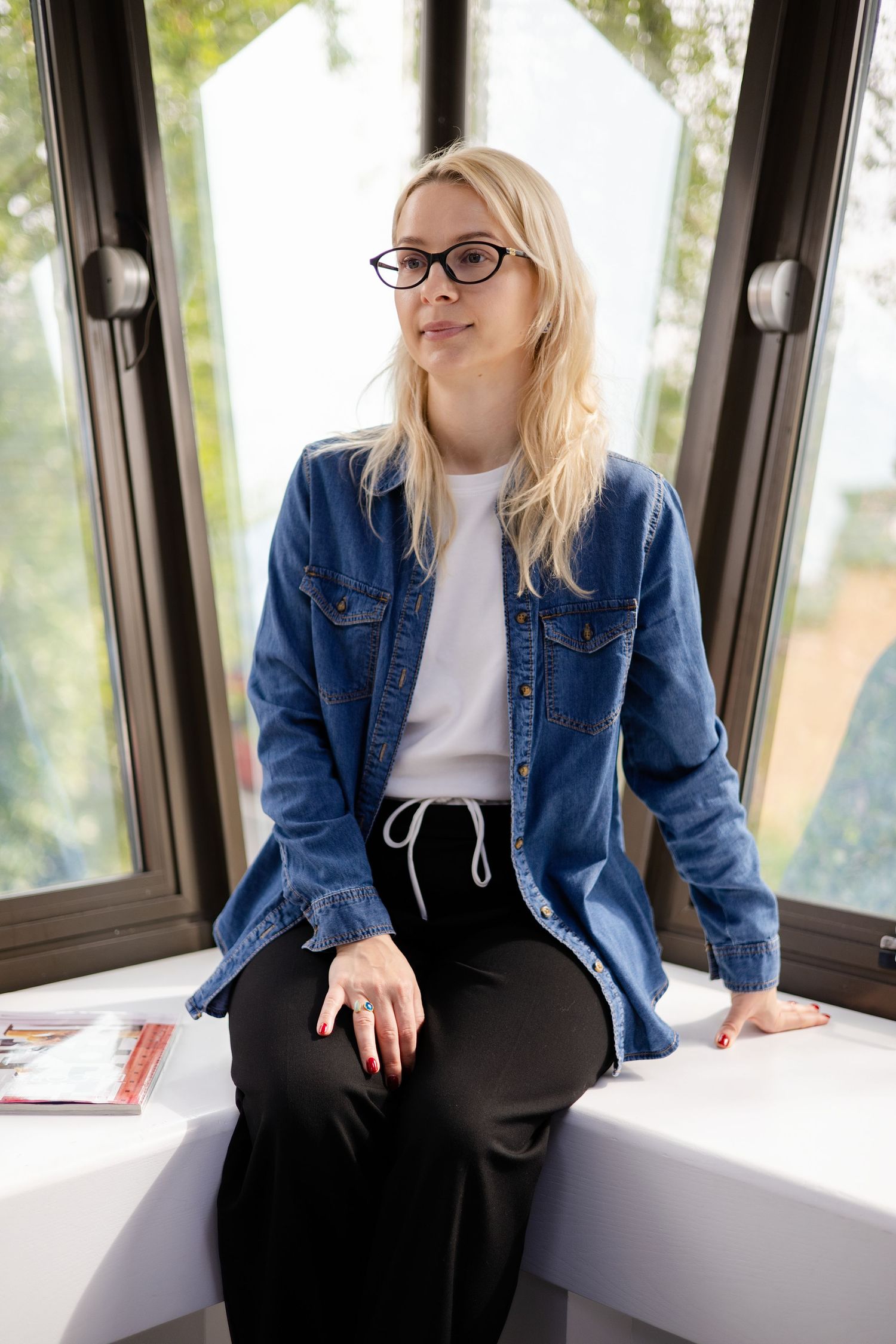 Blonde woman in glasses and denim jacket sitting on windowsill holding magazine in modern space. Brand Photography South London