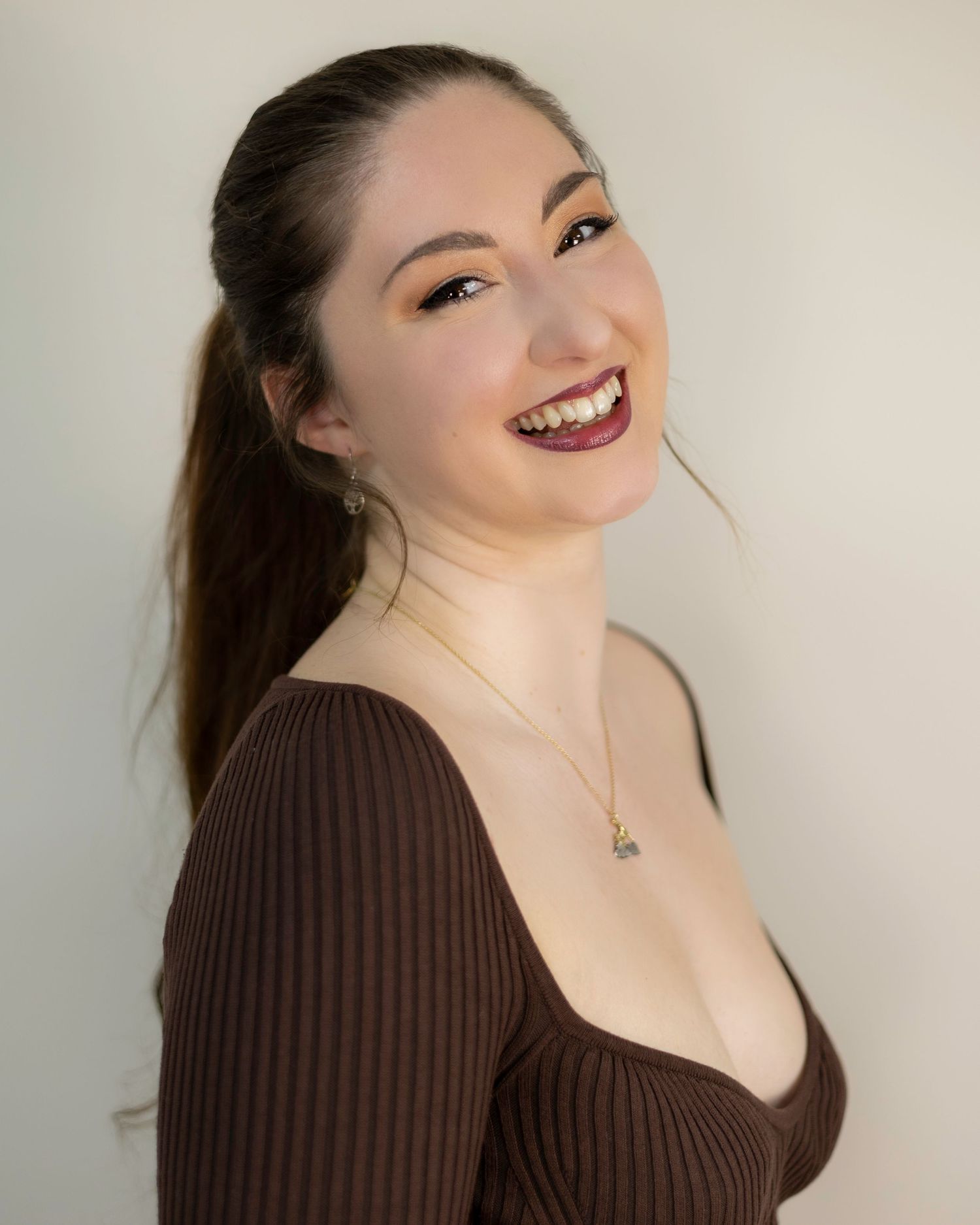 Smiling young woman with dark hair in ponytail wearing brown ribbed top and gold necklace posing against white background. London