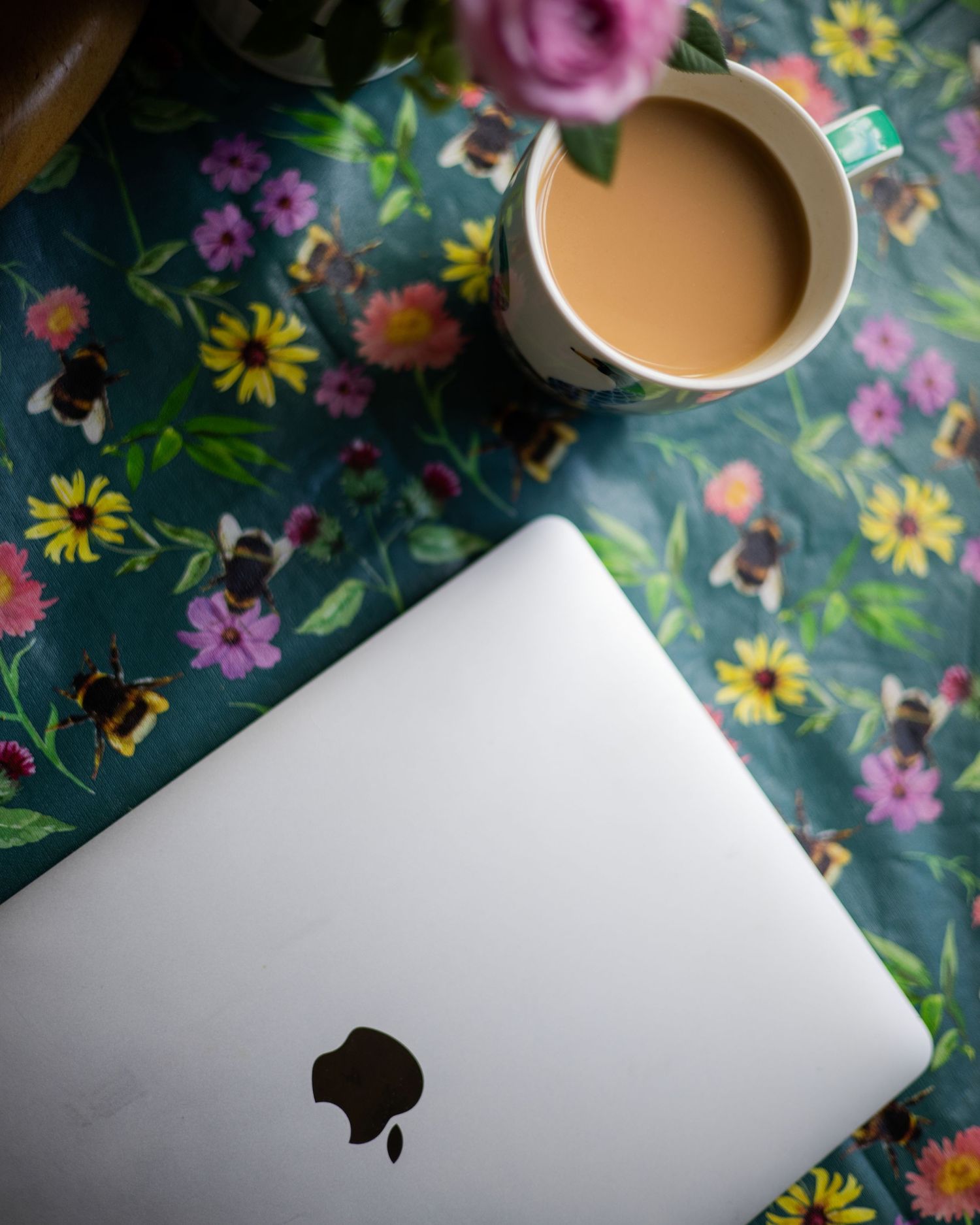 Apple MacBook laptop beside a cup of tea on a floral bee-print tablecloth with pink and yellow flowers.