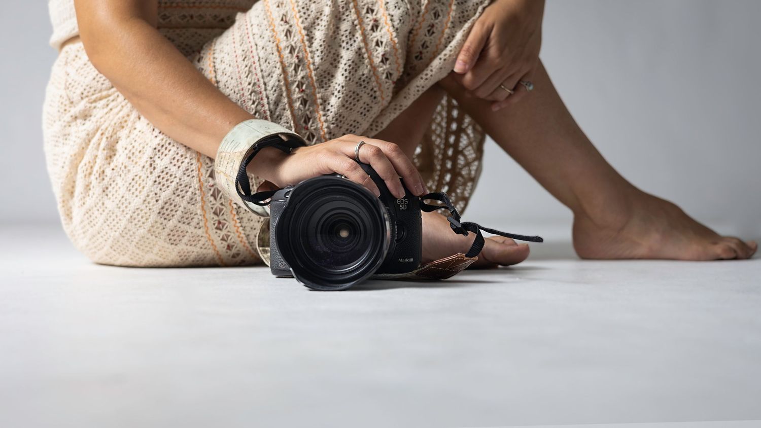 Close-up of woman in cream crochet dress sitting barefoot on white floor holding a DSLR camera. Blanes, Spain