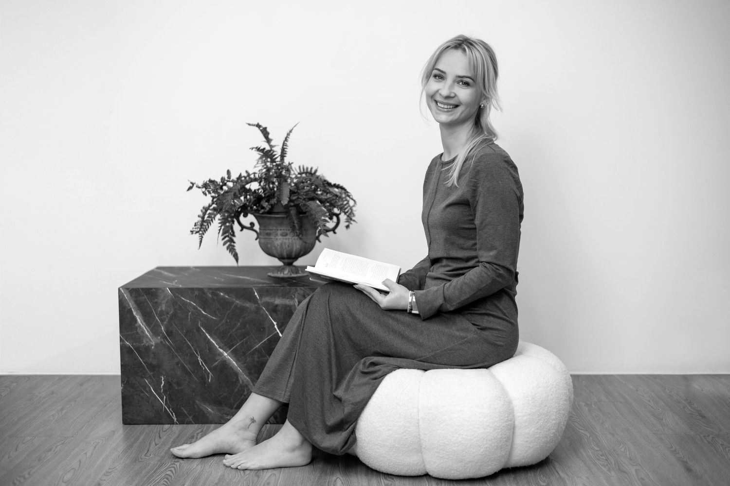 Smiling blonde woman sitting on white pouf holding book beside marble table with plant, black and white photo. London
