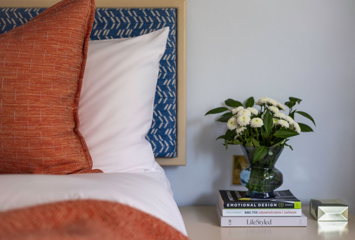 Cozy bedroom nightstand with white flowers, stacked books beside orange and blue bedding. Brand Photography South London
