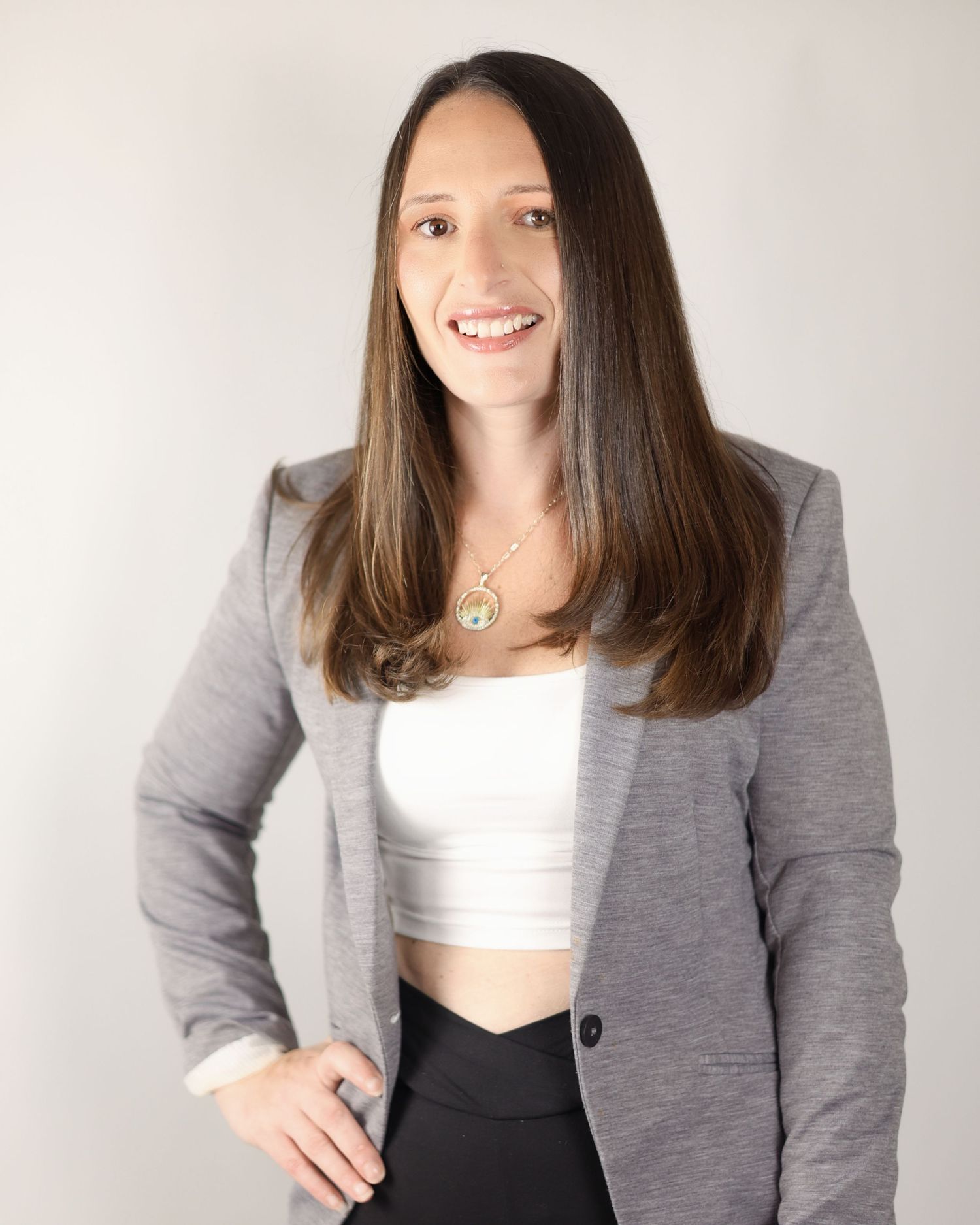 Smiling brunette woman in gray blazer and white crop top with gold pendant necklace headshot. South London Baker
