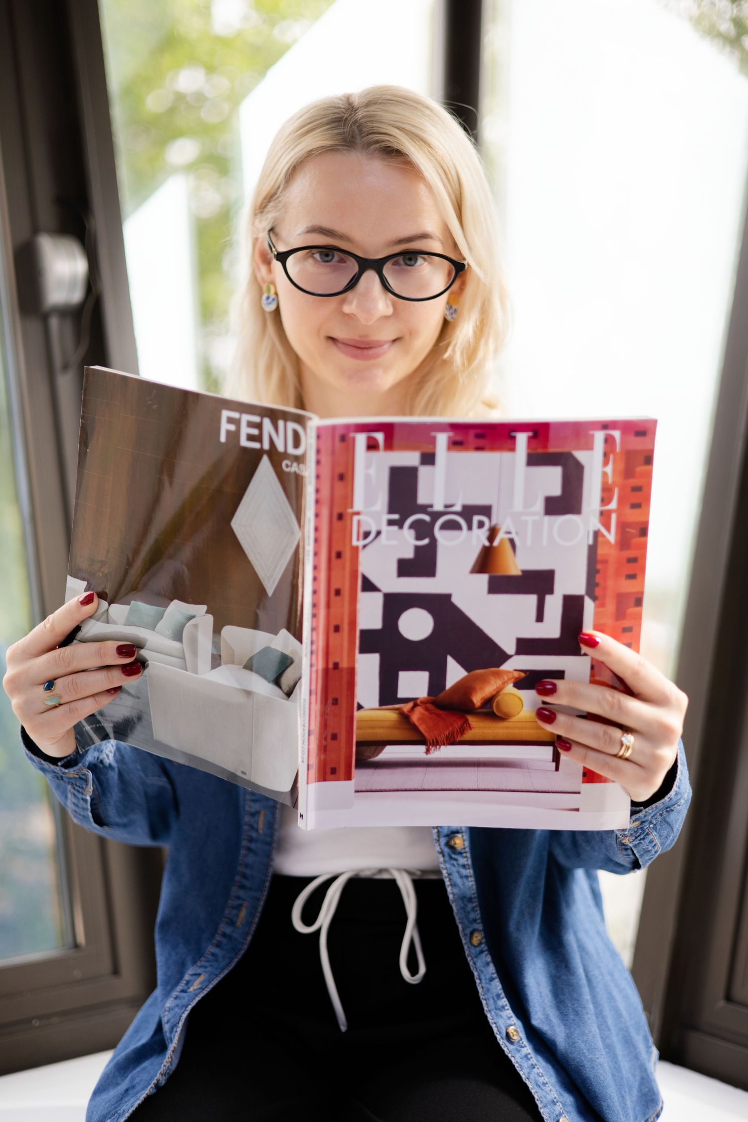 Blonde woman in glasses holding Elle Decoration and Fendi magazines sitting by window. Brand Photography South London