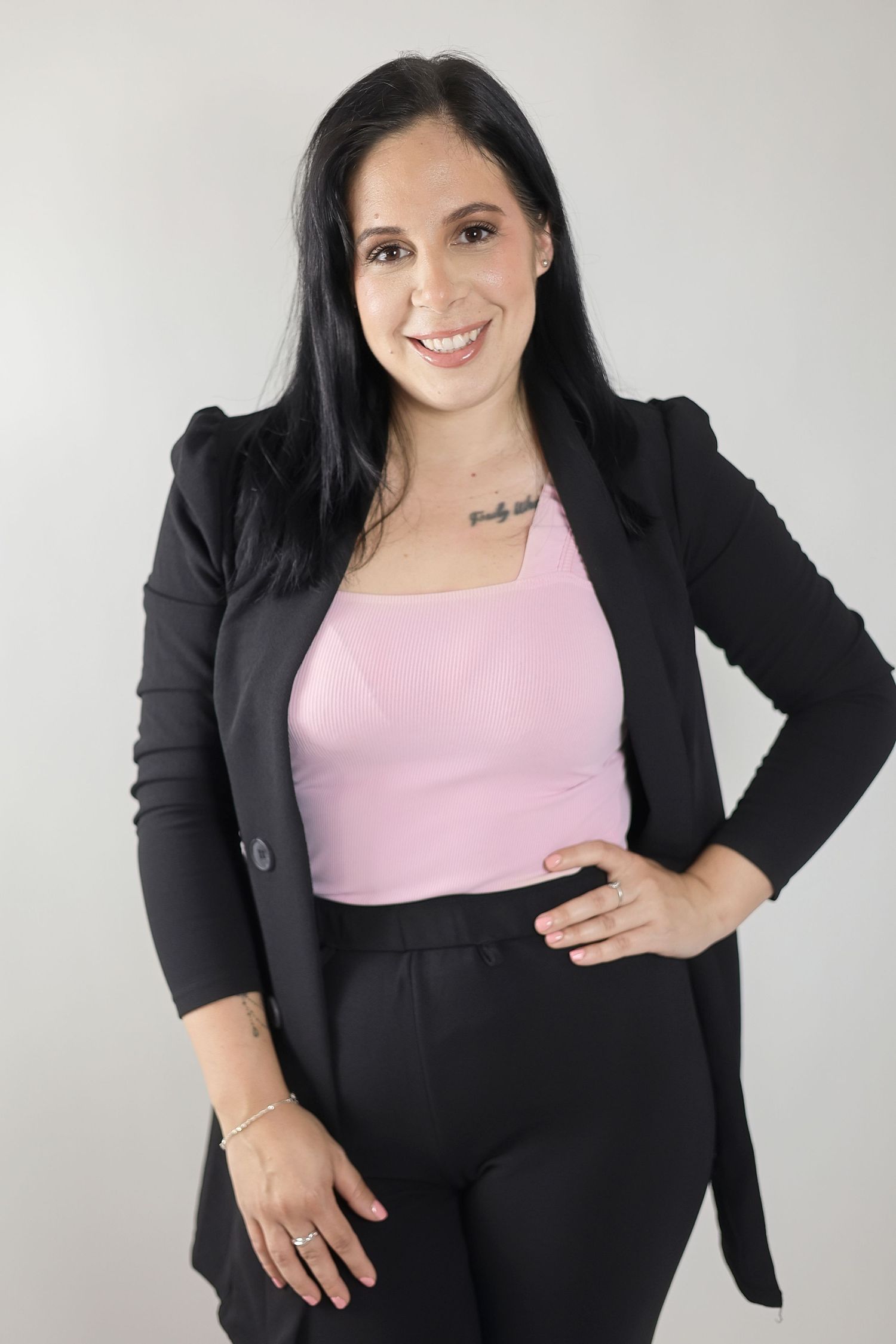 Woman in black blazer and pink top smiling confidently with hand on hip against gray background. Brand Photography South London