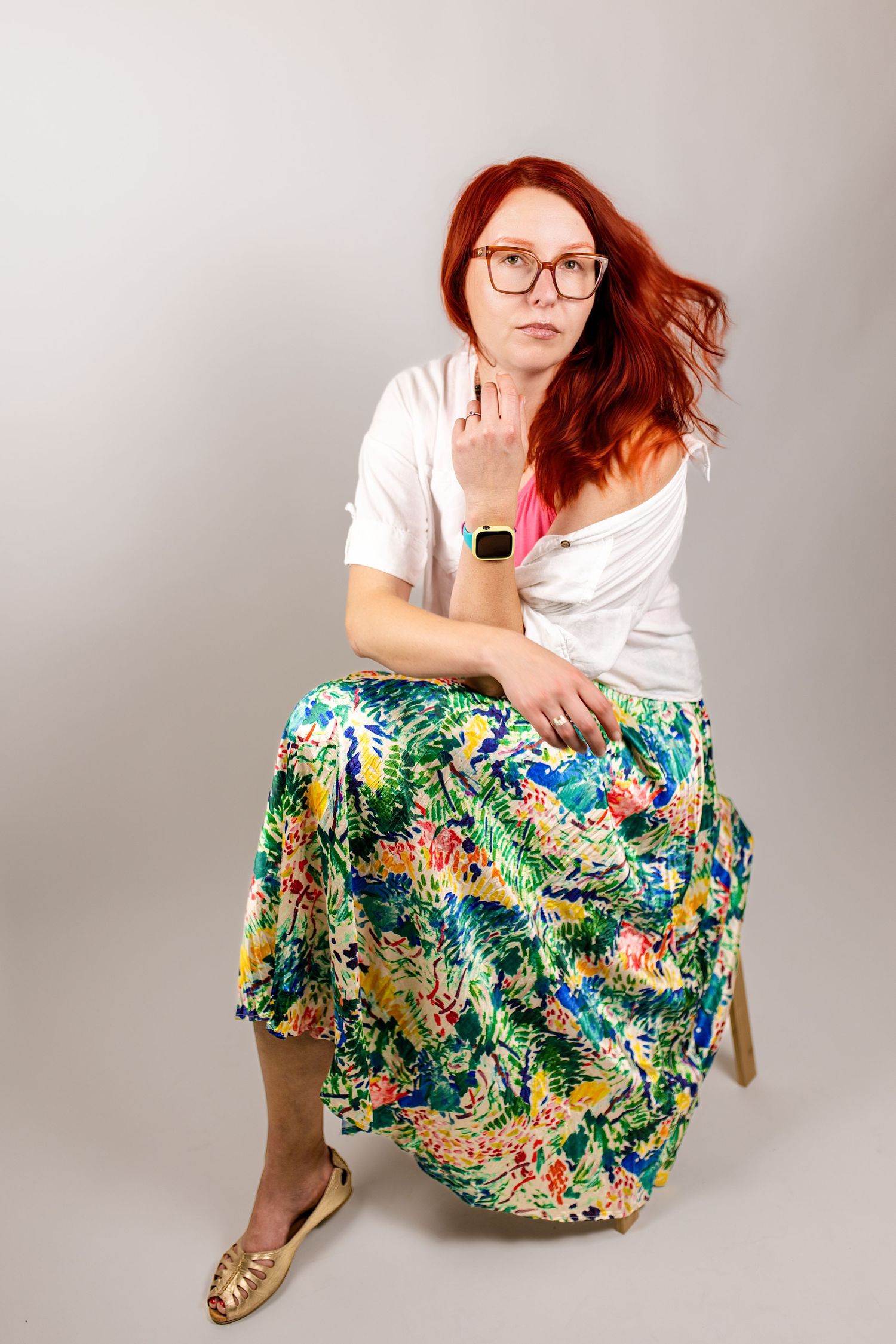 Red-haired woman in glasses wearing floral skirt and white top sitting on stool in studio. Coulsdon