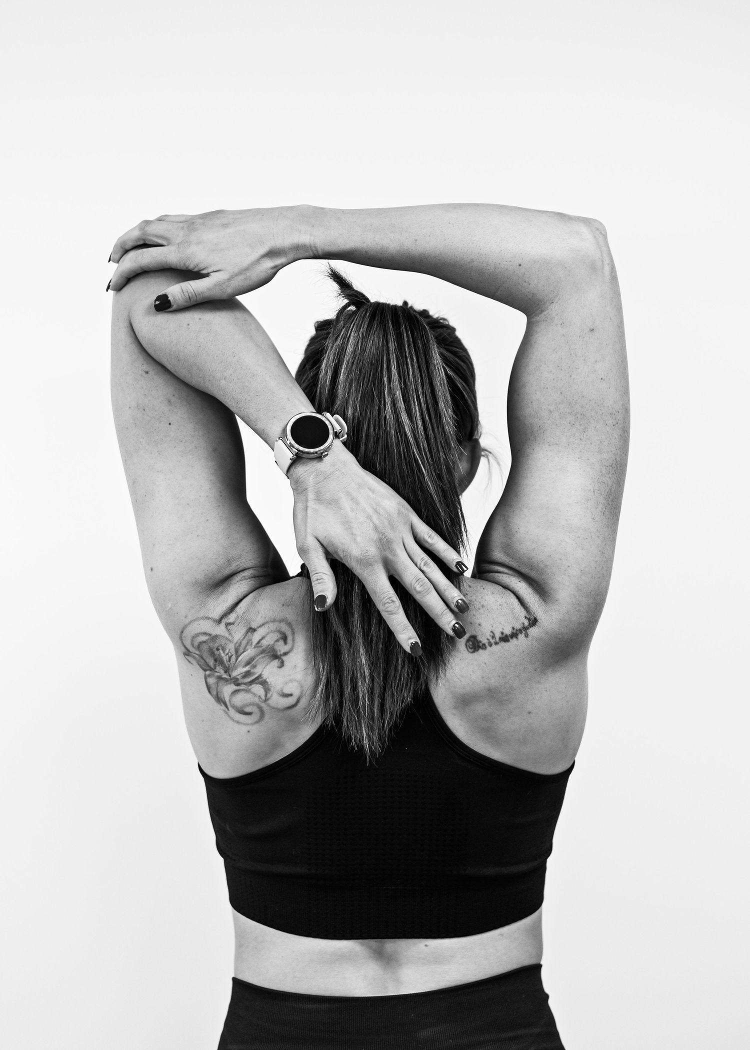 Black and white rear view of tattooed woman stretching arms above head wearing black sports bra. Brand Photography South London
