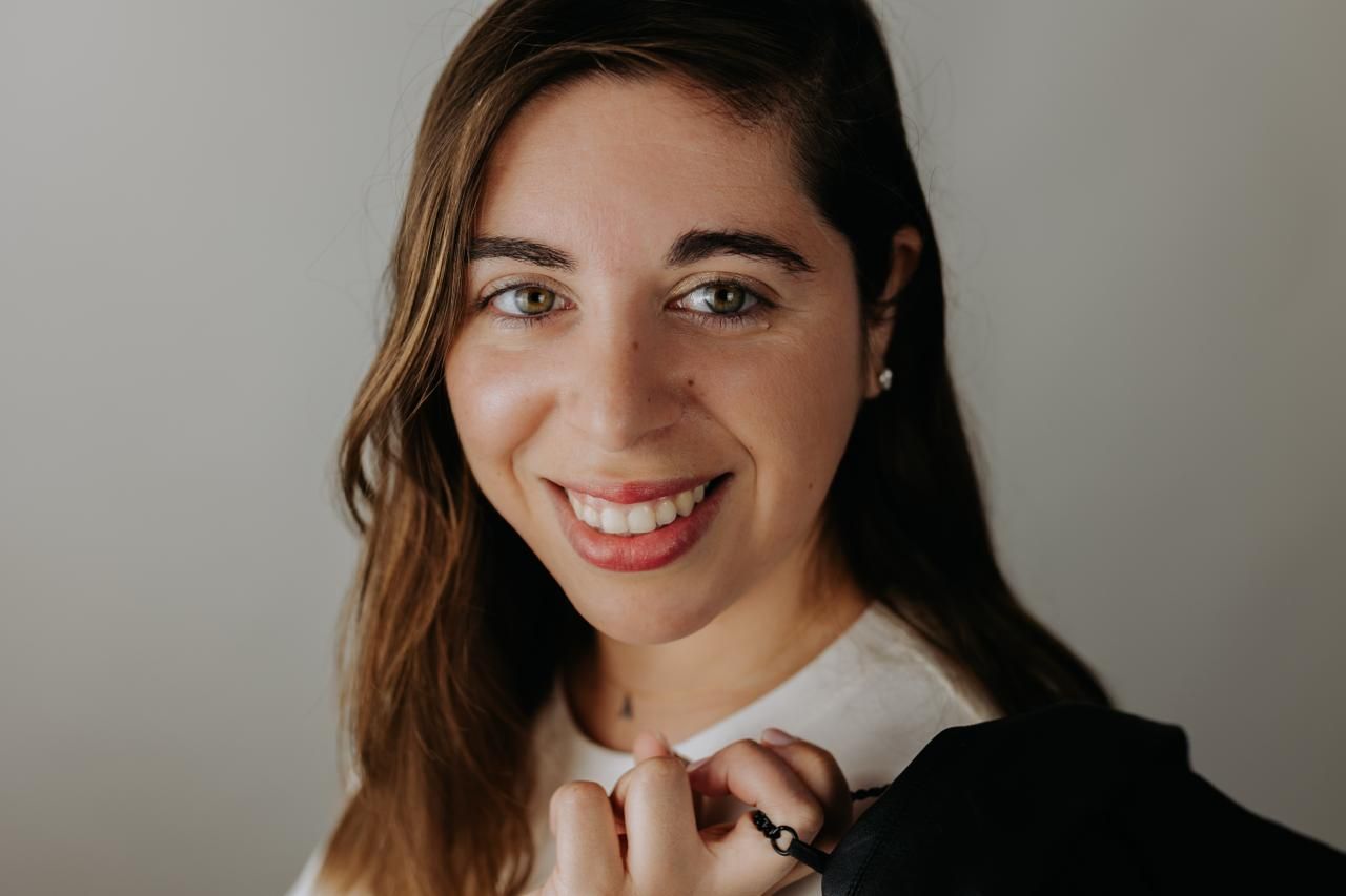 Close-up portrait of smiling brunette woman in black blazer against light gray background. South London