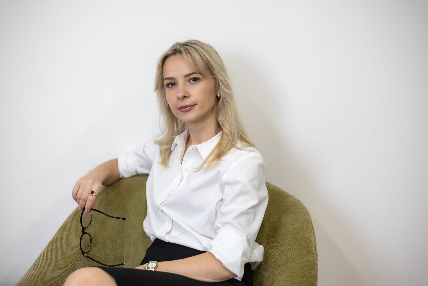Young blonde woman in white shirt sitting in green chair holding glasses against white wall.