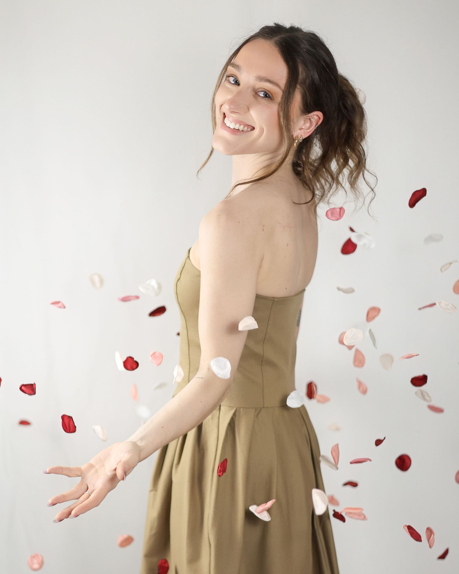 Smiling woman in a strapless olive dress surrounded by falling rose petals in a studio setting. Brand Photography South London