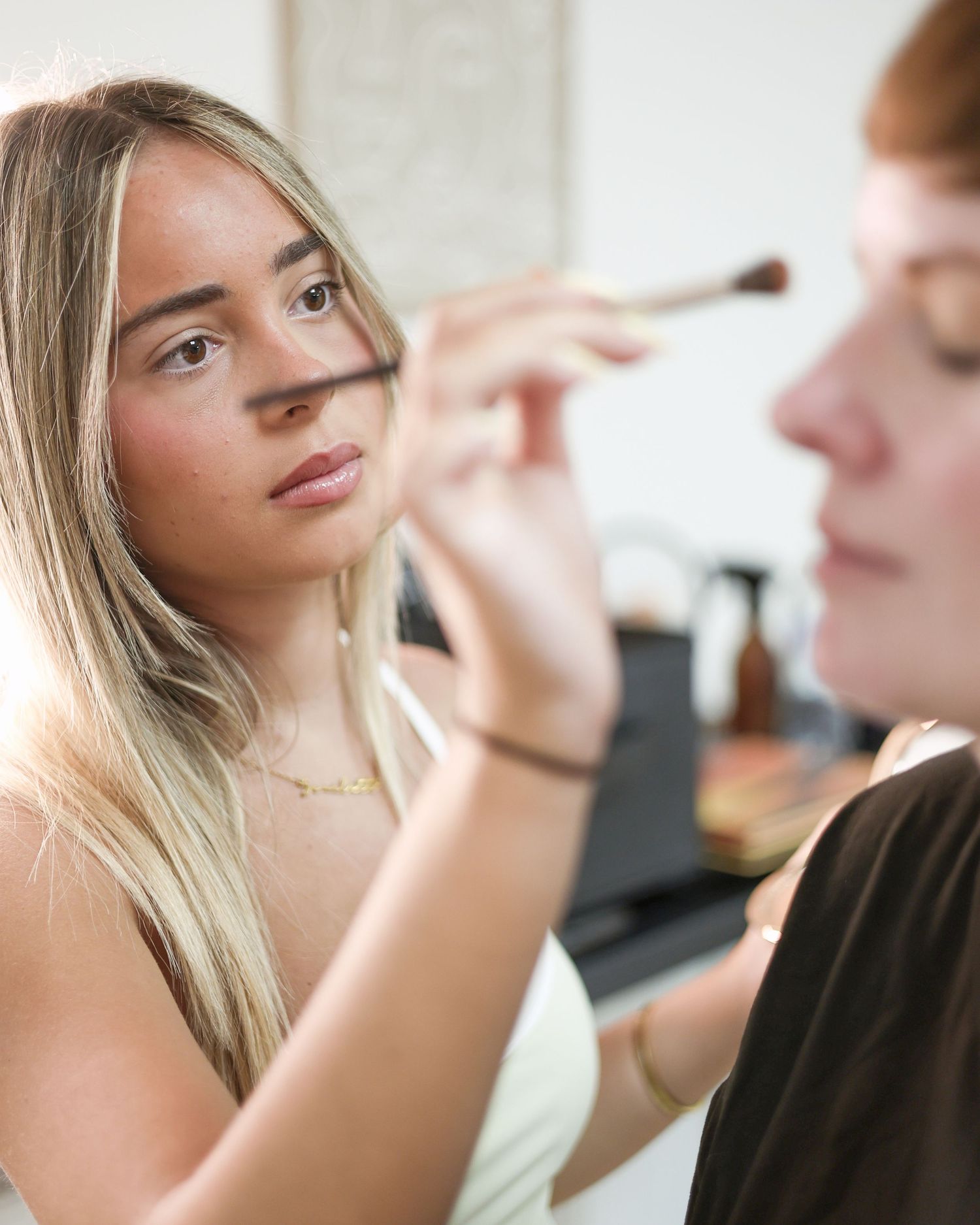 Young blonde makeup artist applying blush to redhead client with brush, beauty salon. South London