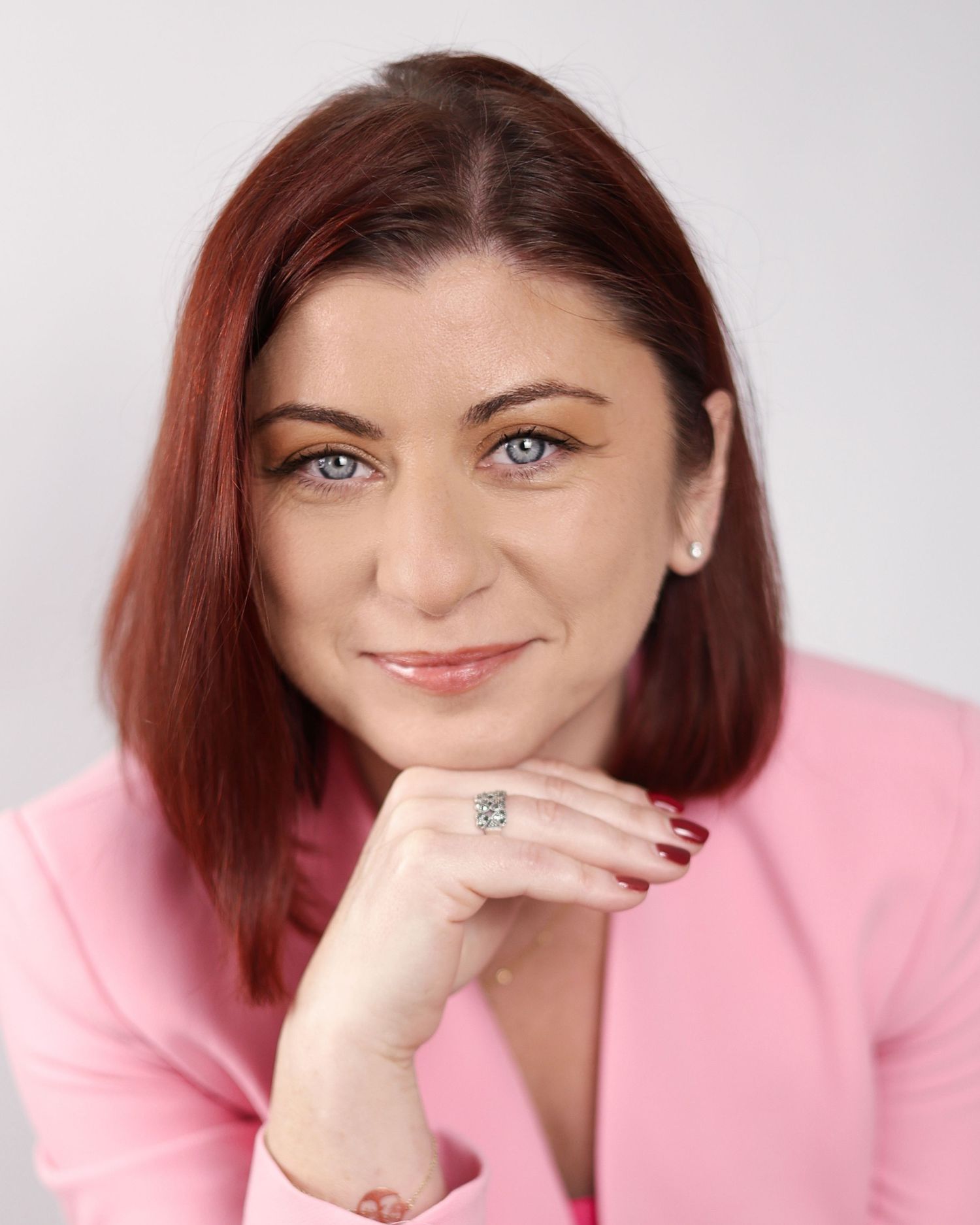 Red-haired woman in pink blazer posing with hand on chin, smiling warmly in professional headshot. Coach in London