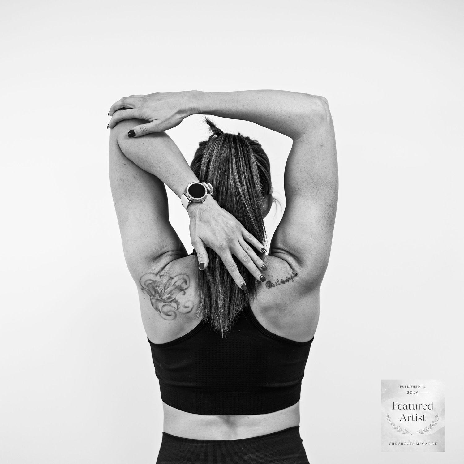 Black and white photo of a woman stretching arms overhead showing back tattoos wearing a sports bra. Brand Photography South London