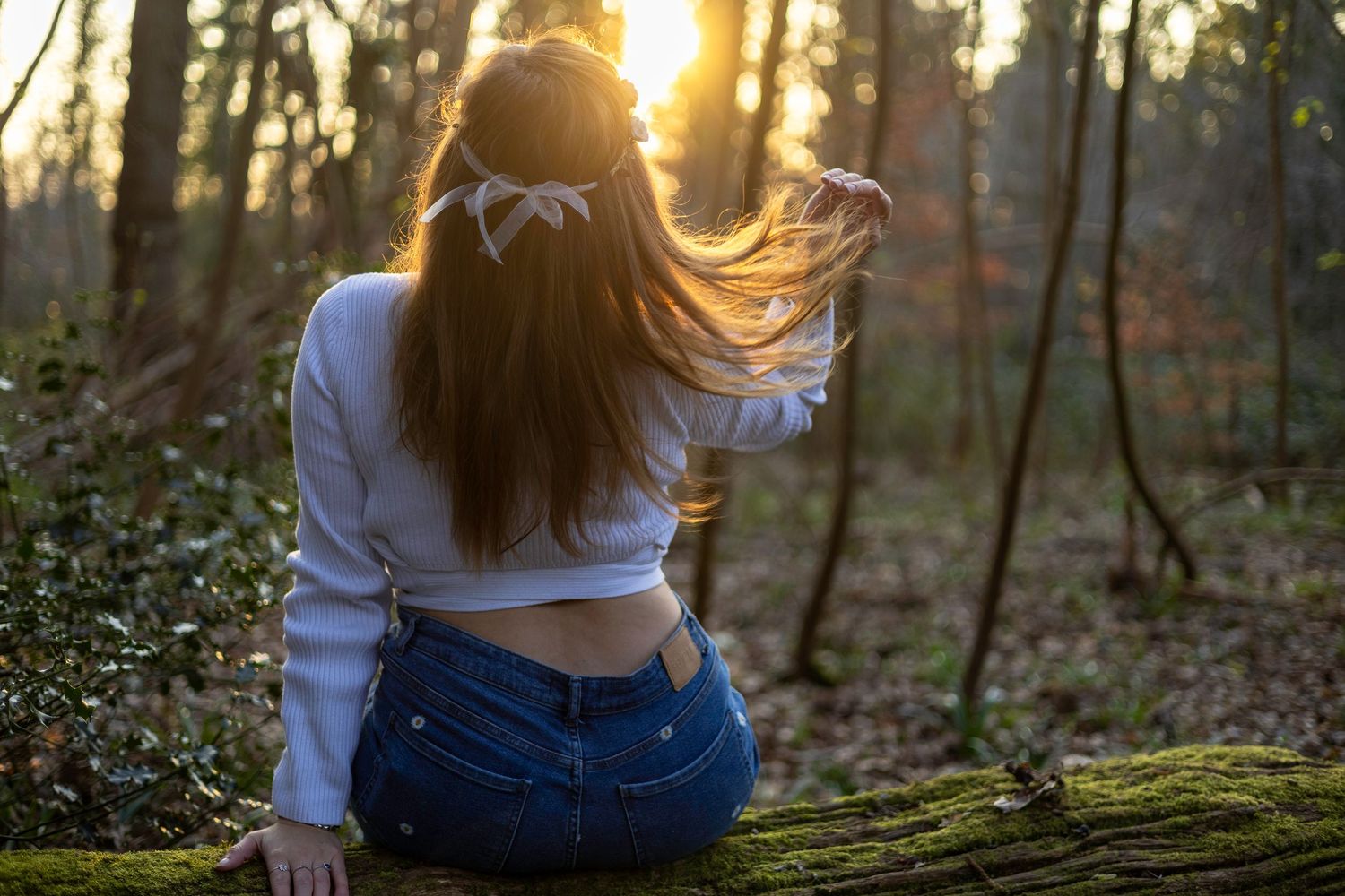 Young woman with long hair and white bow sits on mossy log in sunlit forest wearing crop top and jeans. South Croydon Brand Photography