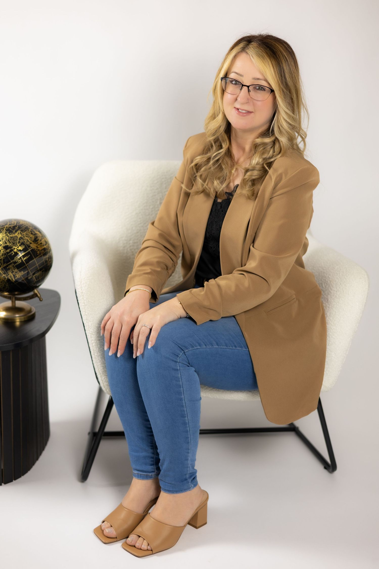 Blonde woman wearing glasses and tan blazer sitting in white chair next to decorative globe on table. South London travel agent