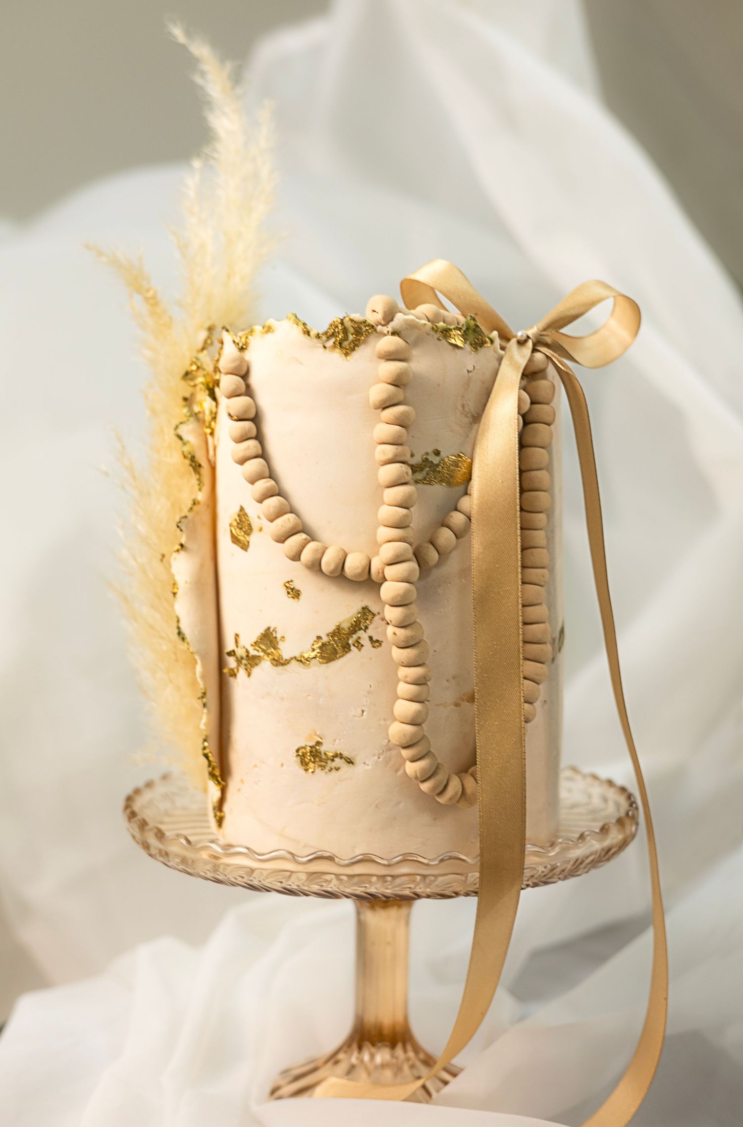 Elegant ivory wedding cake with gold leaf accents, pearl beading, ribbon bow, and dried pampas grass on a gold cake stand. South London