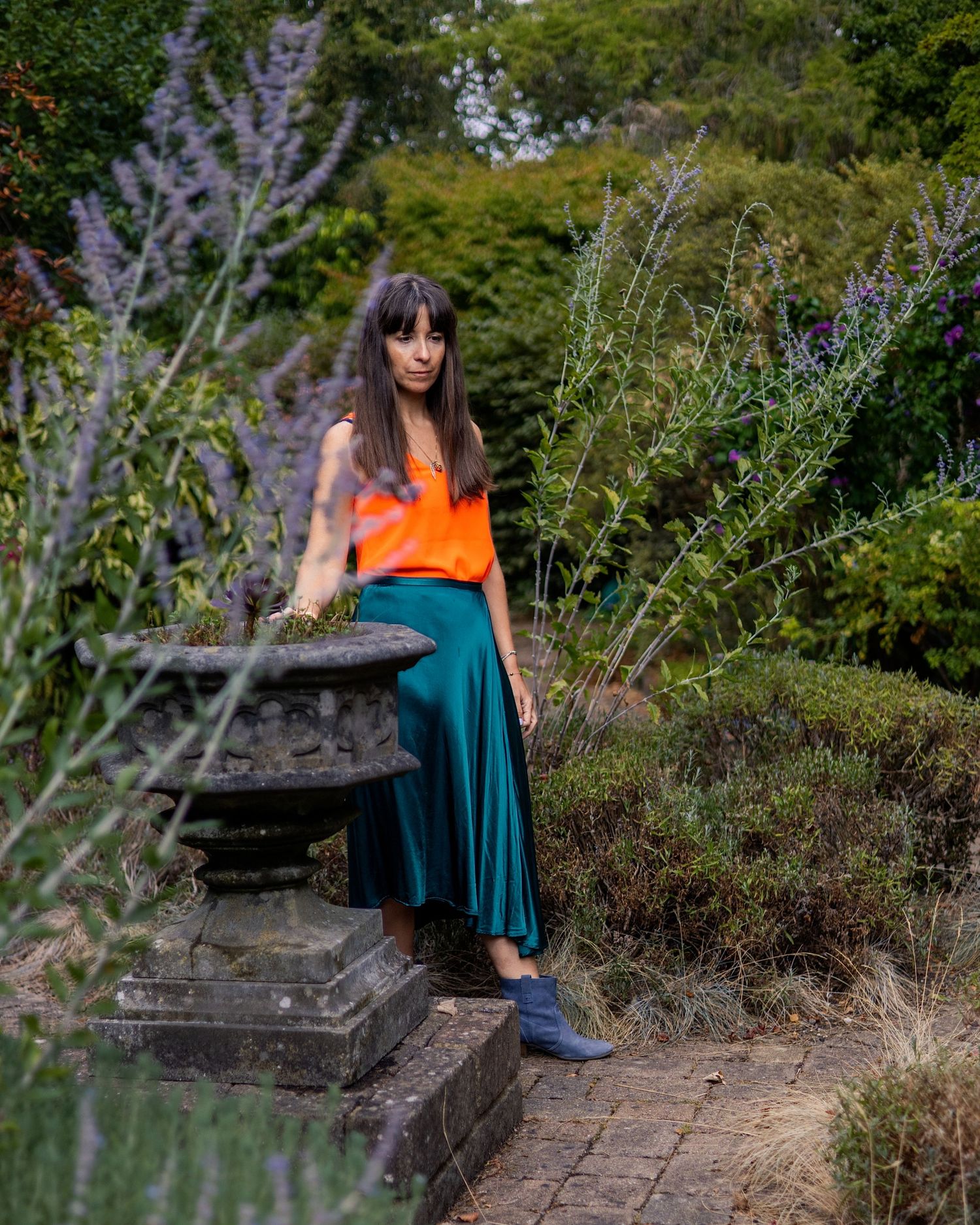 Woman in orange top and teal skirt poses by stone lantern in lush garden setting.