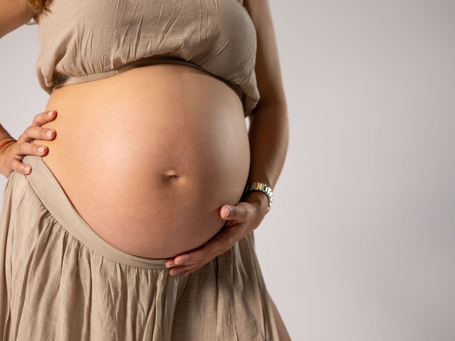 Close-up of a pregnant woman in beige outfit cradling her bare baby bump against a light background. Blanes, Spain