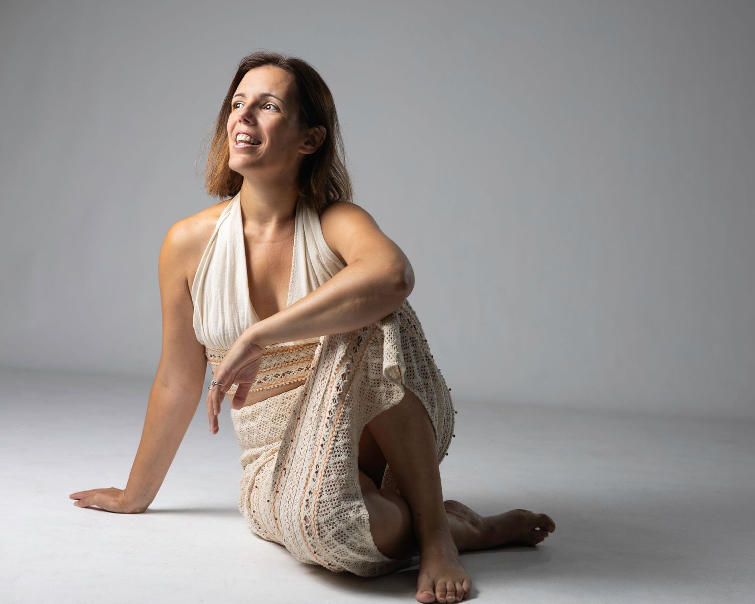 Smiling woman in cream halter dress sitting barefoot on floor, leaning on knee against grey background. Blanes, Spain