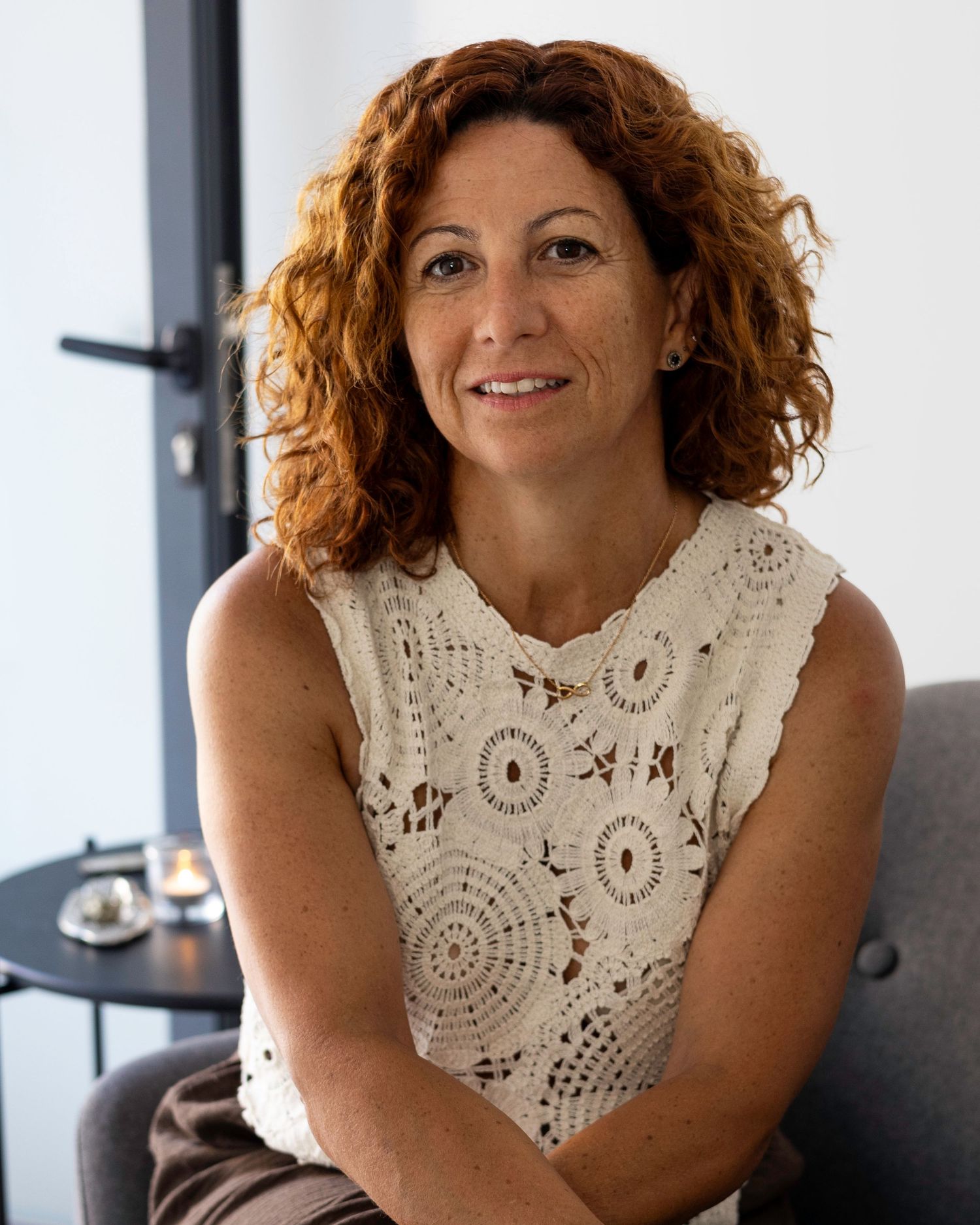 Smiling woman with curly red hair in white crochet top seated indoors. Lloret de Mar, Spain