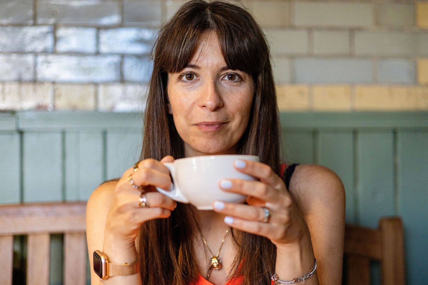 Woman holding white coffee cup at cafe table, wearing red top and gold jewelry, with teal wall background.