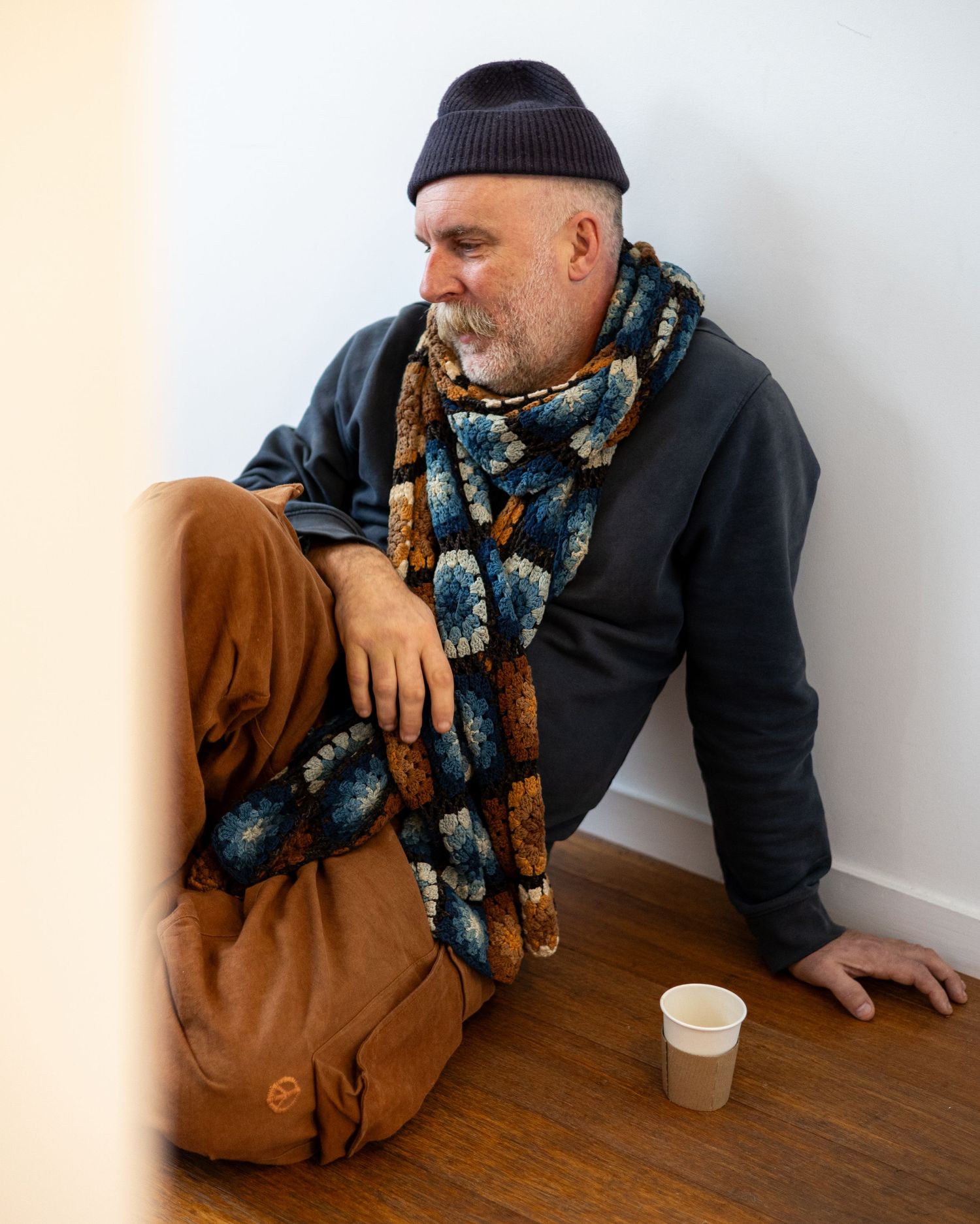 Bearded man in beanie hat and colorful crochet scarf sitting on wooden floor with coffee cup and brown bag. Dulcie Founder, London