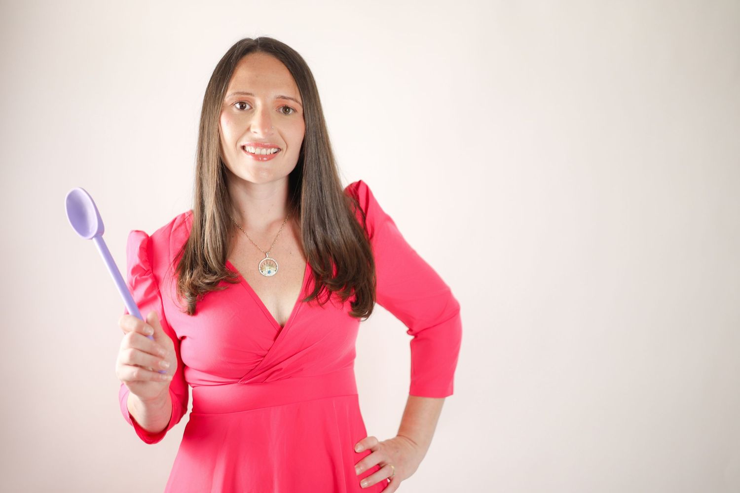 Smiling brunette woman in pink wrap dress holding purple spoon against white background. Baker in Croydon
