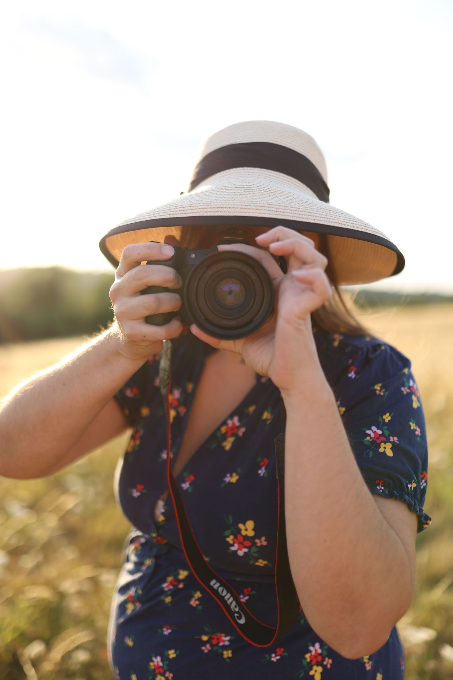 Photographer in floral dress and straw hat taking photo with Canon camera in sunny field. South London
