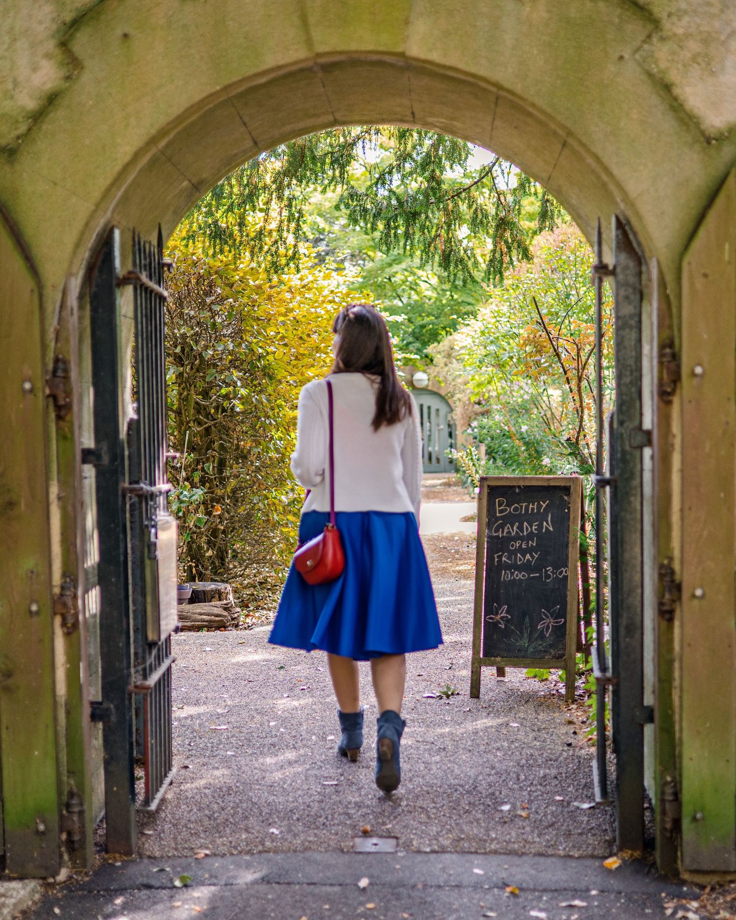 Woman in blue skirt and white top walks through stone archway into a lush garden near a chalkboard sign.