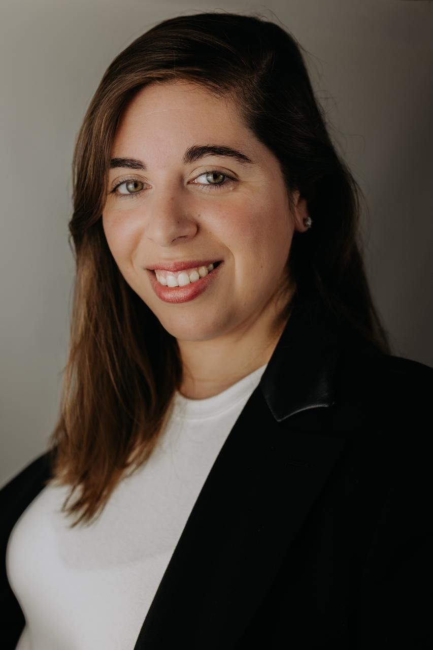 Professional headshot of smiling brunette woman in black blazer and white top against gray background. South London