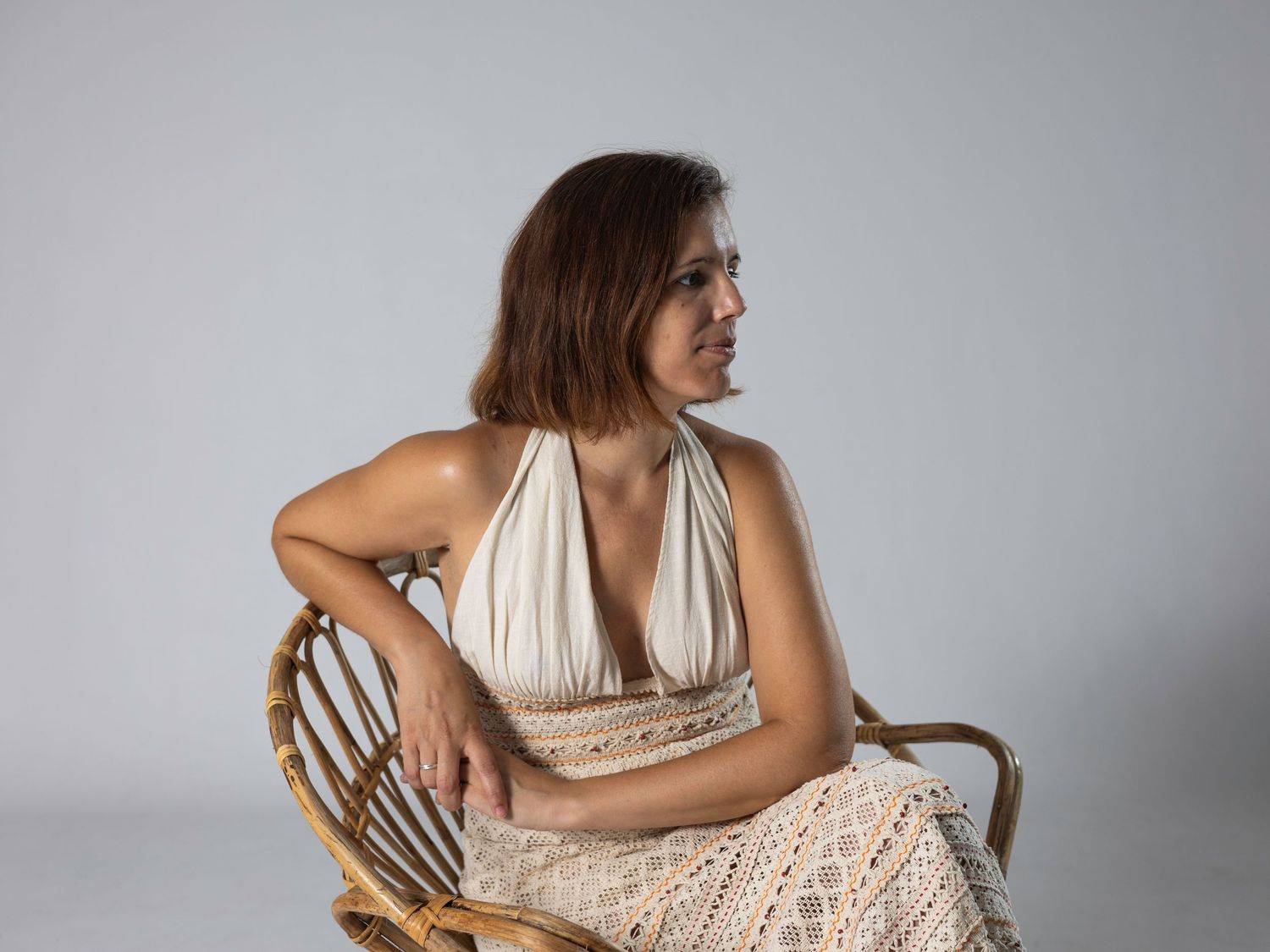Woman in cream halter dress sitting in rattan chair looking sideways against light grey background.