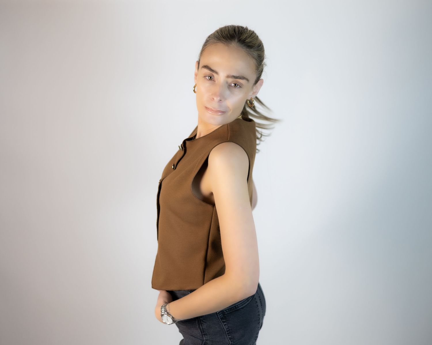 Young woman in brown sleeveless top and jeans posing sideways against a light gray background. South London