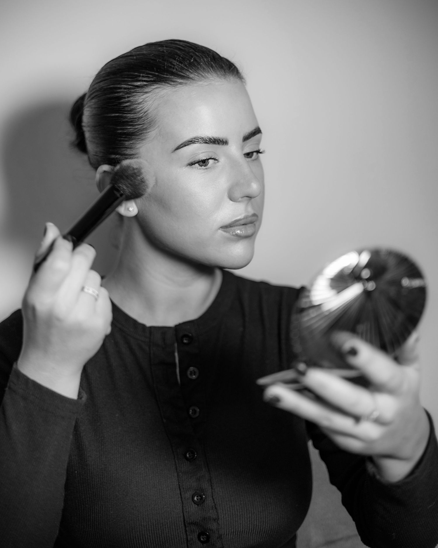Black and white photo of woman applying blush with brush while looking into compact mirror. Make up artist in London