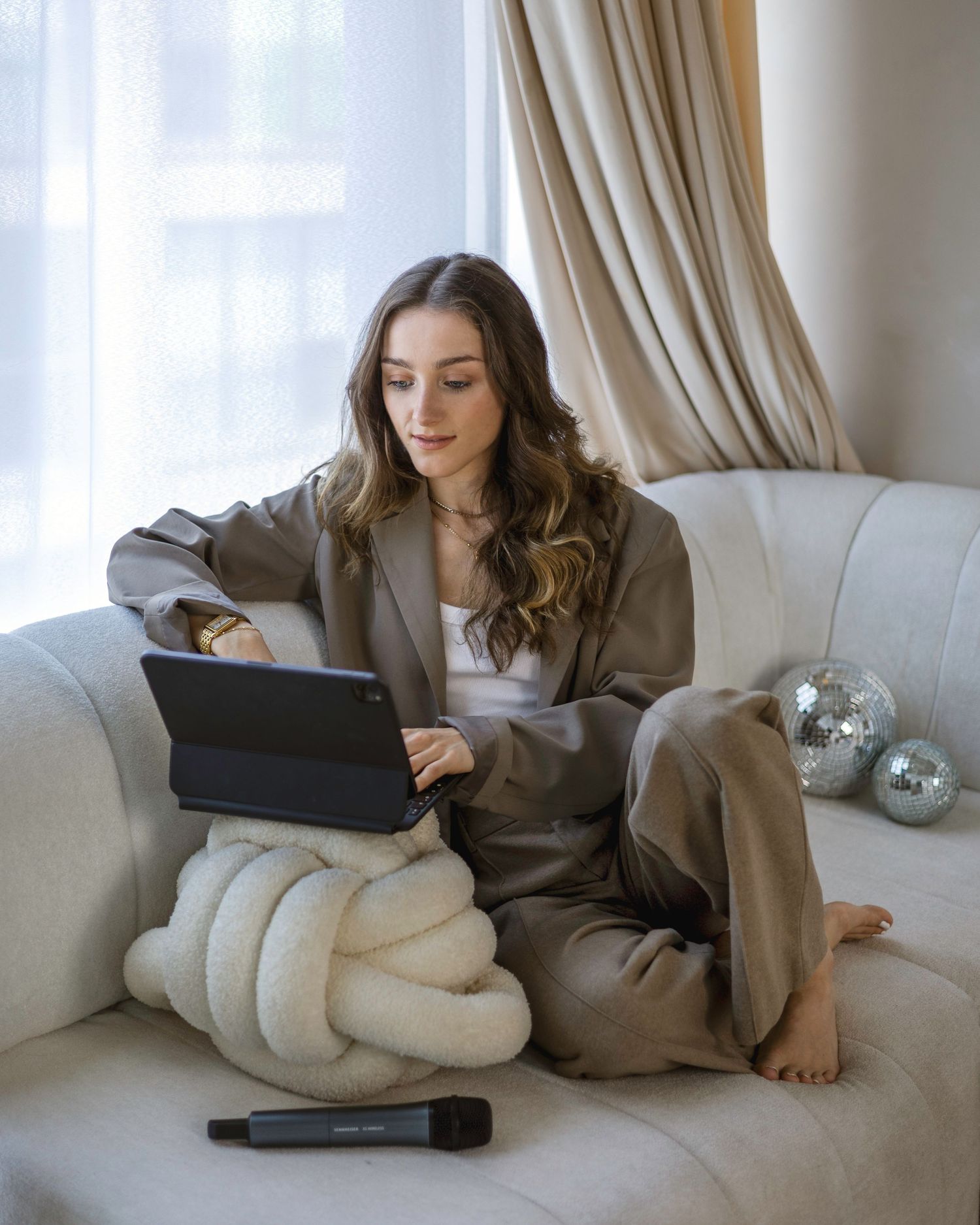 Woman in gray/brown suit sitting on white sofa using tablet, with microphone and disco balls nearby. London