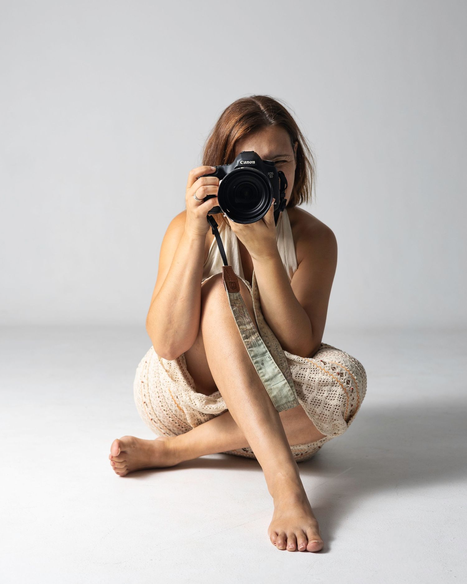 Woman sitting on floor holding Canon camera up to her face, wearing cream crochet dress, barefoot on white background. Blanes, Spain